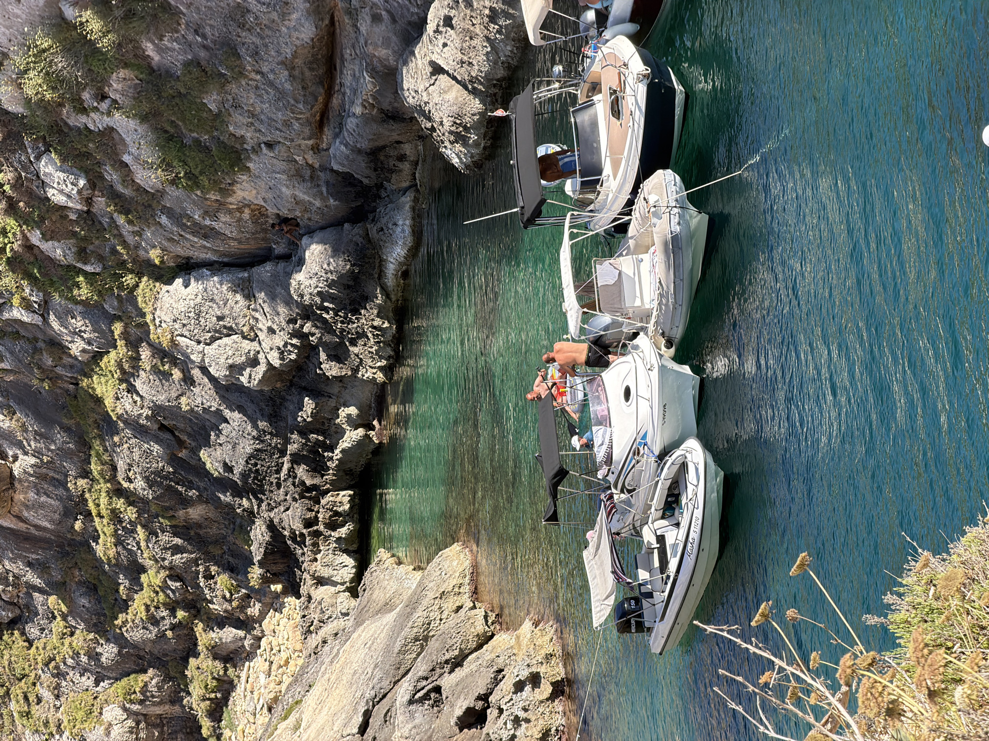 The image captures a sun-drenched scene in the coastal town of Xlendi, Malta, likely within a natural cove or bay. Several boats are anchored in the clear, turquoise water. In the foreground, two white speedboats are positioned close to each other, with a larger boat with a dark hull and a black canopy behind them. A person wearing a white hat is visible in the center speedboat, and two men are standing in the rear speedboat. The water is calm, reflecting the surrounding rugged, rocky cliffs that rise steeply from the water's edge. These cliffs are sparsely covered with vegetation. The overall impression is one of a serene and inviting Mediterranean setting.