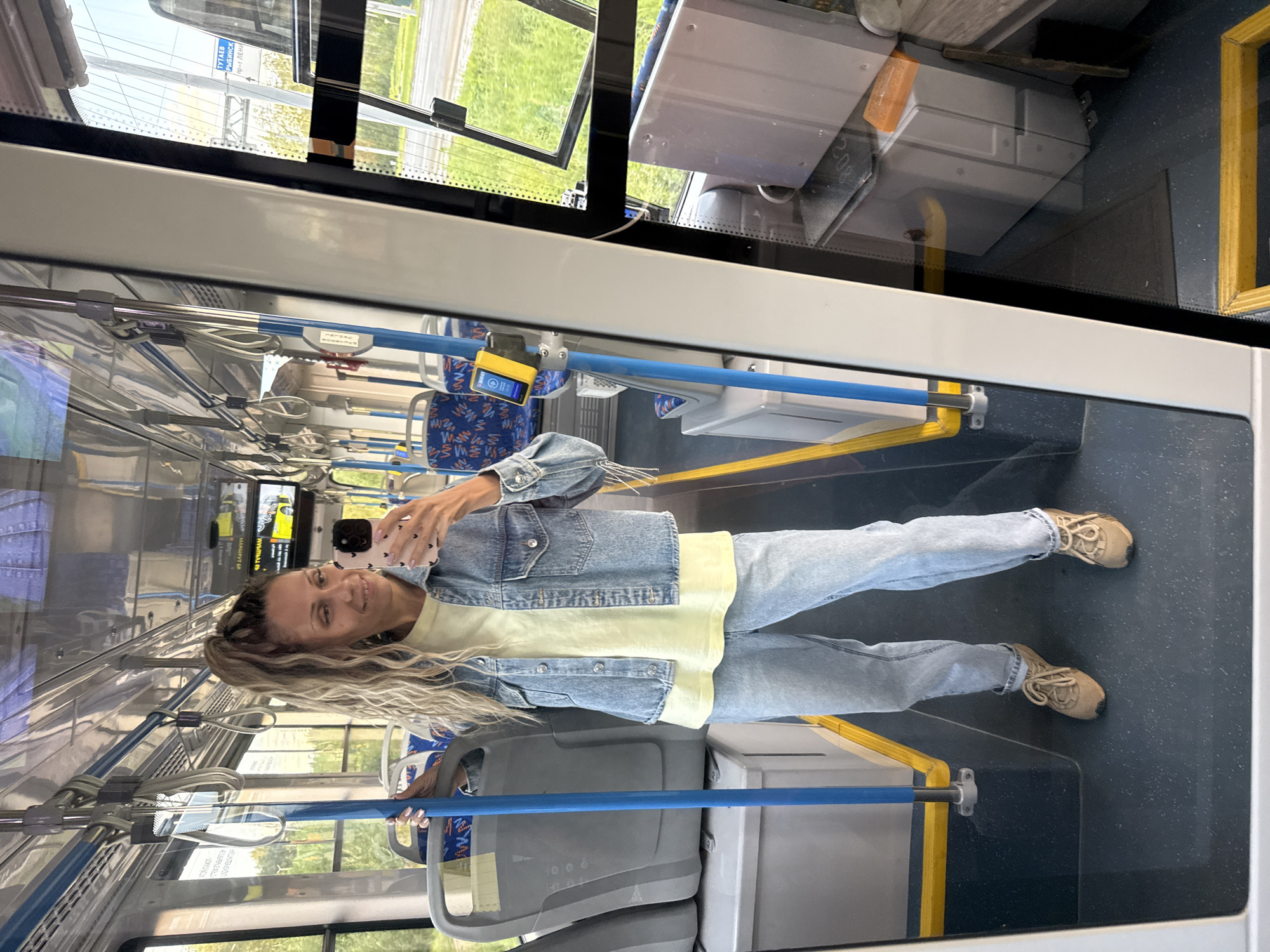 A young woman is taking a selfie in a bus. She is wearing a light blue denim jacket over a yellow t-shirt and light blue jeans, with tan sneakers. Her blonde hair is in a high ponytail. The bus is empty except for her. The interior of the bus is visible, with blue and gray seats and metal poles. The woman is holding a pink phone in her right hand. The scene is lit by natural light coming through the windows. The location is identified as Rusanovo, Russia, based on a sign visible through the window.