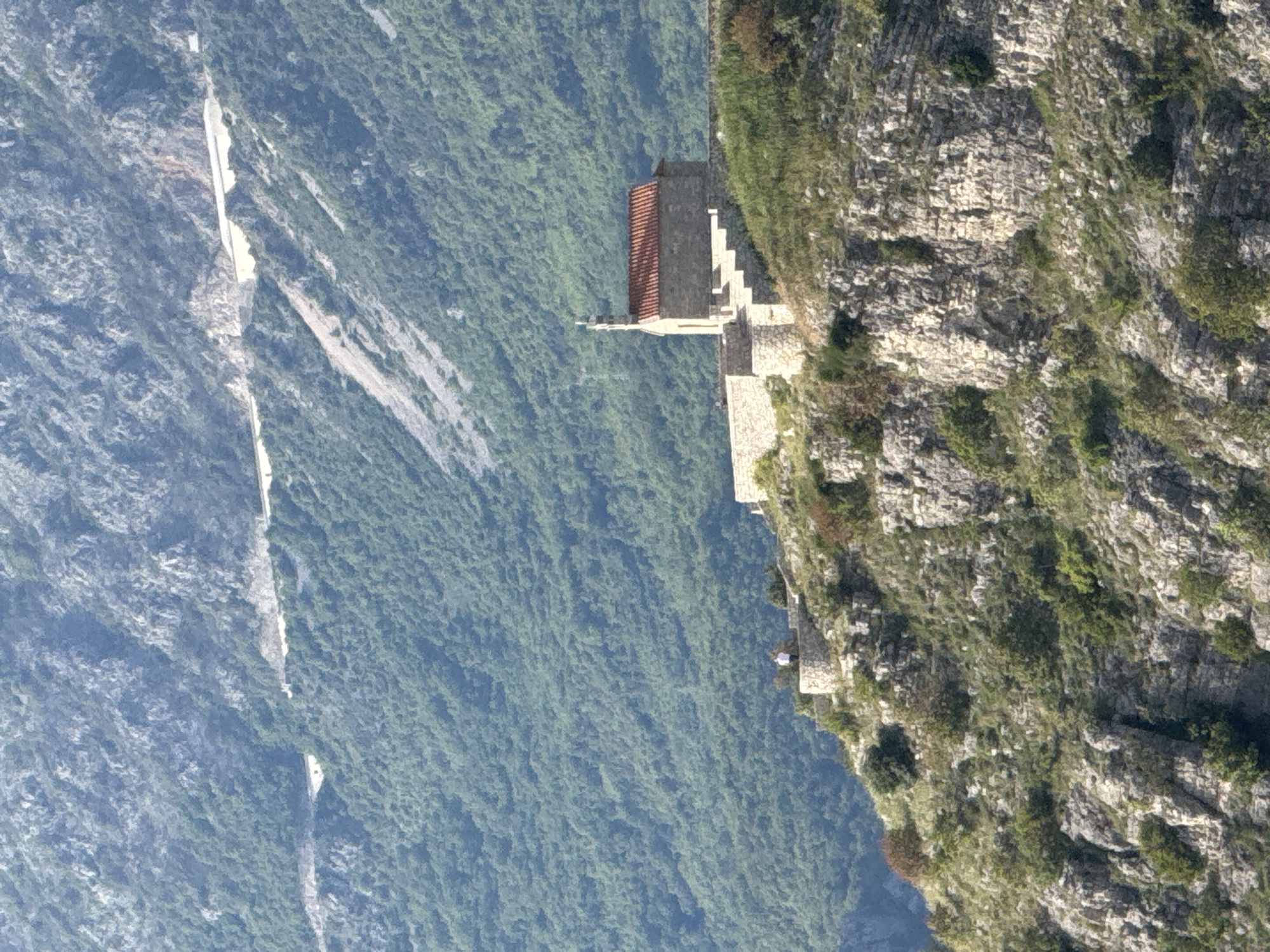 The image shows a cliffside church in Sveti Stefan, Montenegro. The church has a red-tiled roof and is built on a rocky promontory. A staircase leads up to the church. A lush, green forest covers the area behind the church, and a road can be seen in the distance. The sky is overcast.