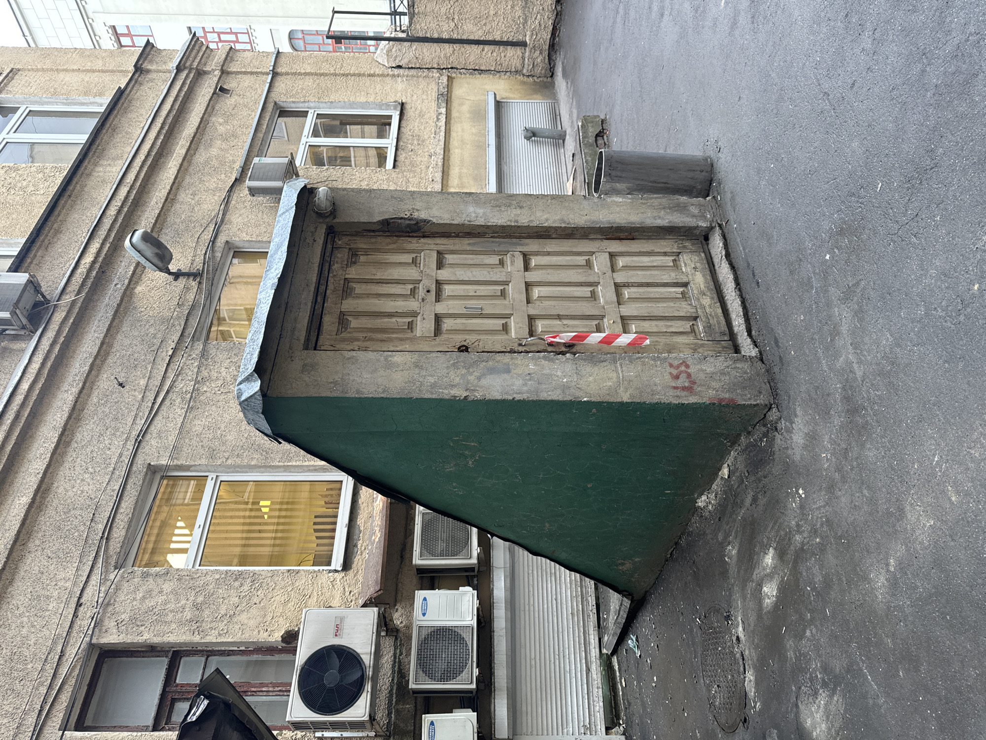 An exterior shot depicts a weathered building in Moscow, Russia, angled towards the right, beside a concrete ground surface. The building is an older structure with multiple windows, some covered by blinds. Several air conditioning units are attached to the building's facade. A low, concrete doorway is visible, partially covered by a green painted structure. A red and white striped barrier tape is placed on the top of the concrete doorway. Graffiti is etched into the concrete surface above the doorway, and a metal pipe is seen above the door. The ground surface is asphalt.