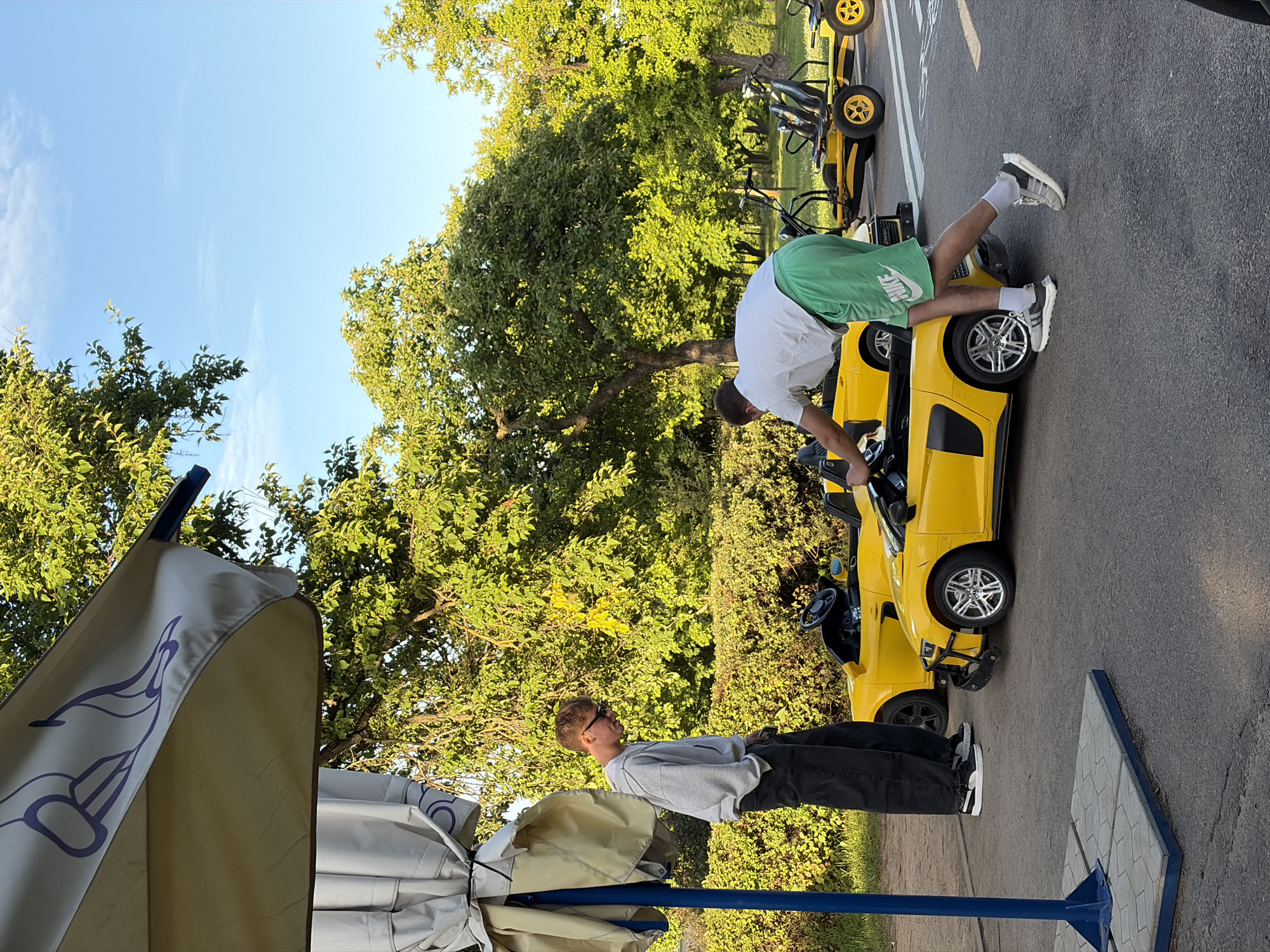 In a park in Minsk, Belarus, two young men stand near a fleet of bright yellow ride-on cars. One man, wearing a grey hoodie and dark pants, stands with his hands in his pockets, looking down at a yellow sports car. The other man, in a white t-shirt and green shorts, leans into a yellow sports car, appearing to adjust something. The cars are parked on an asphalt path, surrounded by lush green trees and bushes under a clear blue sky. A large, pale umbrella with a purple design is partially visible in the foreground, providing shade. Several other yellow ride-on vehicles, including what appear to be go-karts, are parked further in the background. The scene suggests a recreational area designed for children's activities or rentals.