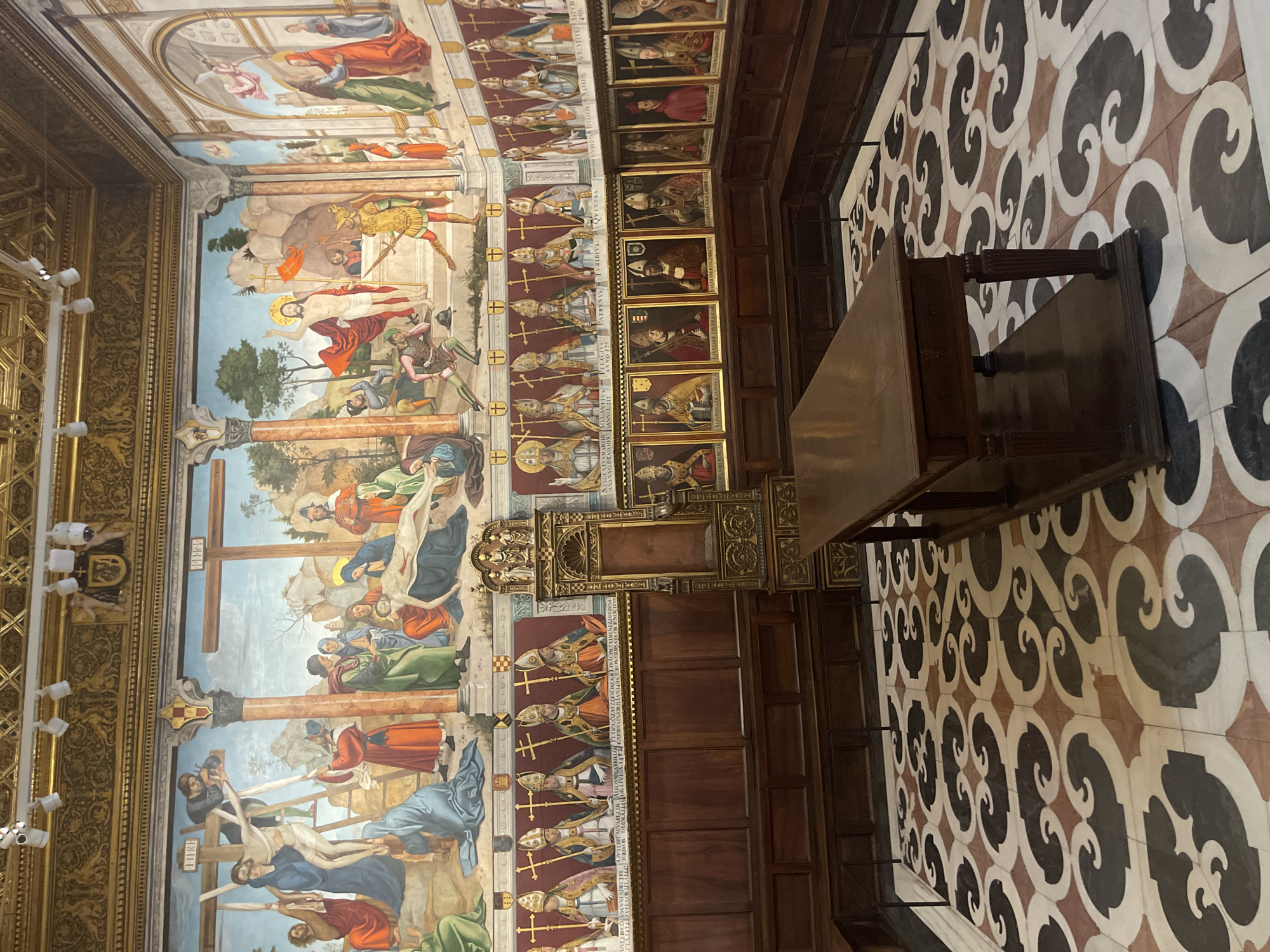 The image shows a richly decorated room in the Primate Cathedral of Saint Mary of Toledo, Spain. The walls are adorned with a series of large frescoes depicting biblical scenes, including the Crucifixion and the Resurrection of Jesus. Below the frescoes, a series of portraits of bishops and cardinals in their religious attire are displayed in ornate frames. A large, gilded wooden throne sits centrally against the wall, flanked by wooden choir stalls. In the foreground, a long, dark wooden table with carved legs rests on a floor covered with a distinctive geometric patterned tile. The overall impression is one of grandeur and historical significance, typical of a major cathedral.
