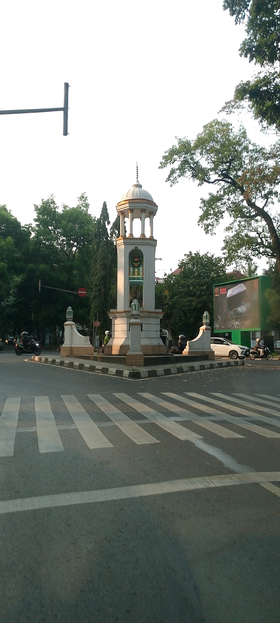 The image shows a street intersection with a clock tower in the center. The clock tower is white with a gold dome and octagonal pillars. It has a clock face visible in the upper section. There are traffic lights, trees, and buildings surrounding the intersection. A crosswalk is prominent in the foreground. Several motorcycles and a white car are present in the scene. A large billboard is visible on the right side of the image. The sky is clear and bright, suggesting it is daytime. This intersection is known as Kilometer Nol or Kilometer Zero, a central landmark in Bandung City, Indonesia.