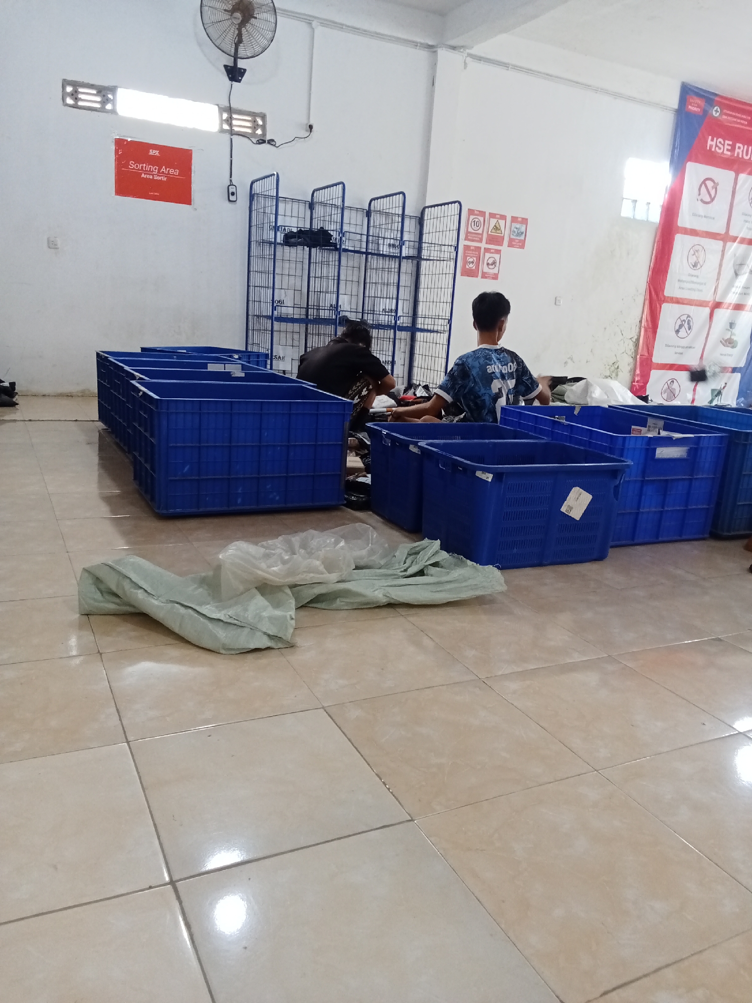 The image depicts an indoor scene that appears to be a sorting or logistics area, possibly within a warehouse or distribution center. The location is identified as Jirak, Indonesia, according to the metadata.
In the foreground, there are several large, blue plastic crates, some empty and some appearing to be in the process of being filled or emptied. A light-colored, possibly fabric, material is draped on the floor in front of the crates, partially obscuring the tiled flooring. The floor is composed of large, beige square tiles.
Two individuals are visible in the mid-ground, engaged in activities related to the crates. One person, wearing a dark shirt, is bent over near a crate. Another person, wearing a blue and white patterned shirt, is also working near the crates.
In the background, there are metal shelving units or racks, typically used for storage and organization. On the wall to the left, there is a red sign with white text that reads "SPX Sorting Area Area Sortir". Above this sign, a wall fan is mounted. To the right of the shelving units, a large banner with red and white elements is partially visible, featuring text and various safety icons. The walls are painted white. The overall impression is one of an active workspace focused on handling goods.