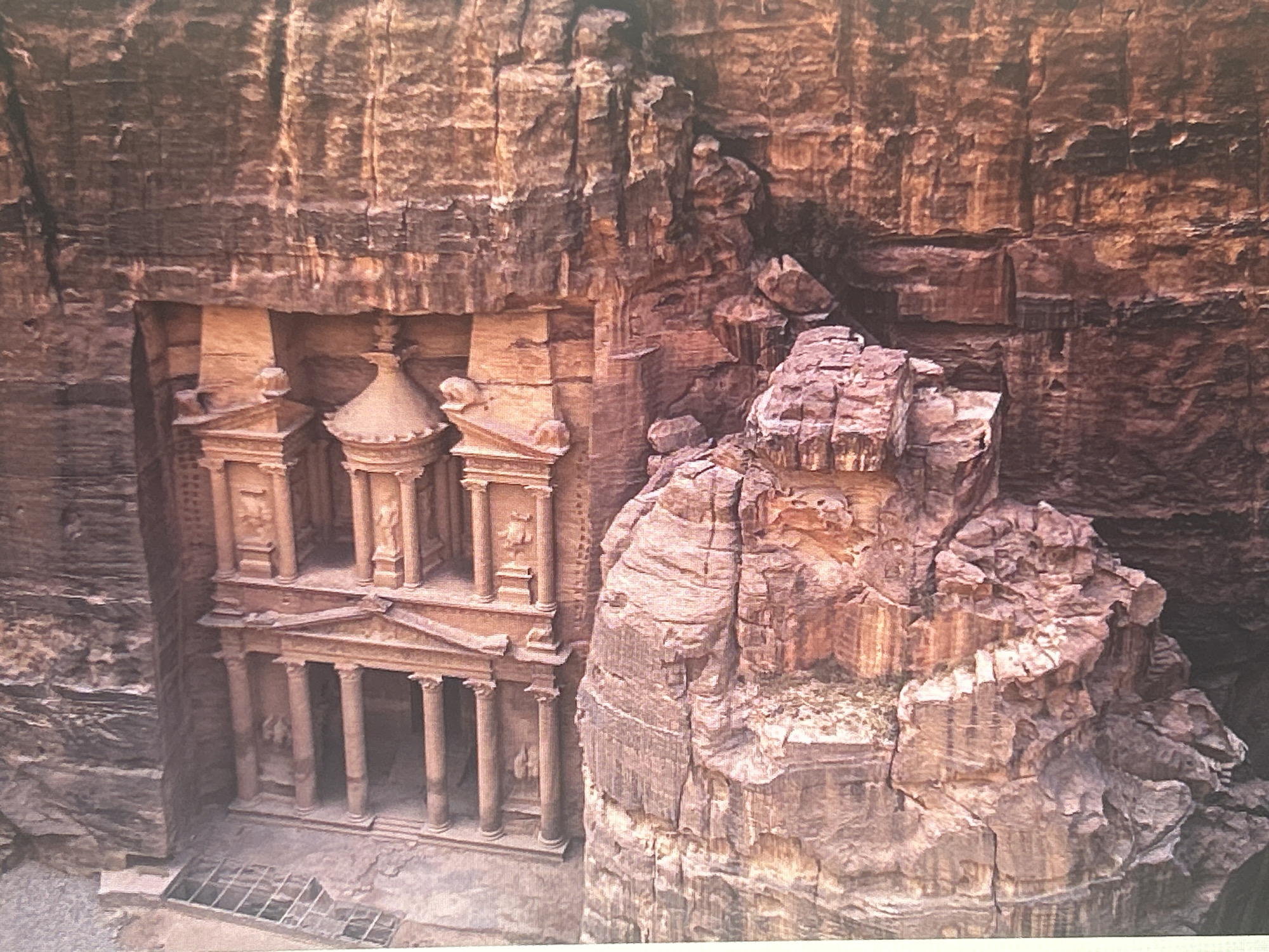 The image displays a wide shot of Al-Khazneh, also known as The Treasury, a famous facade carved directly into a sandstone cliff face in Petra, Jordan. The structure is a grand Hellenistic-style monument with intricate columns, pediments, and sculptures. It is situated within a narrow canyon with towering rock walls on either side. The sandstone exhibits layers of varying colors, ranging from pale pink to deep red and brown, indicating geological strata. The lighting suggests it is daytime, with shadows accentuating the architectural details and the ruggedness of the surrounding rock formations. There are no people visible in the image. The environment is arid and rocky. The location is NOT Sofia, Bulgaria.