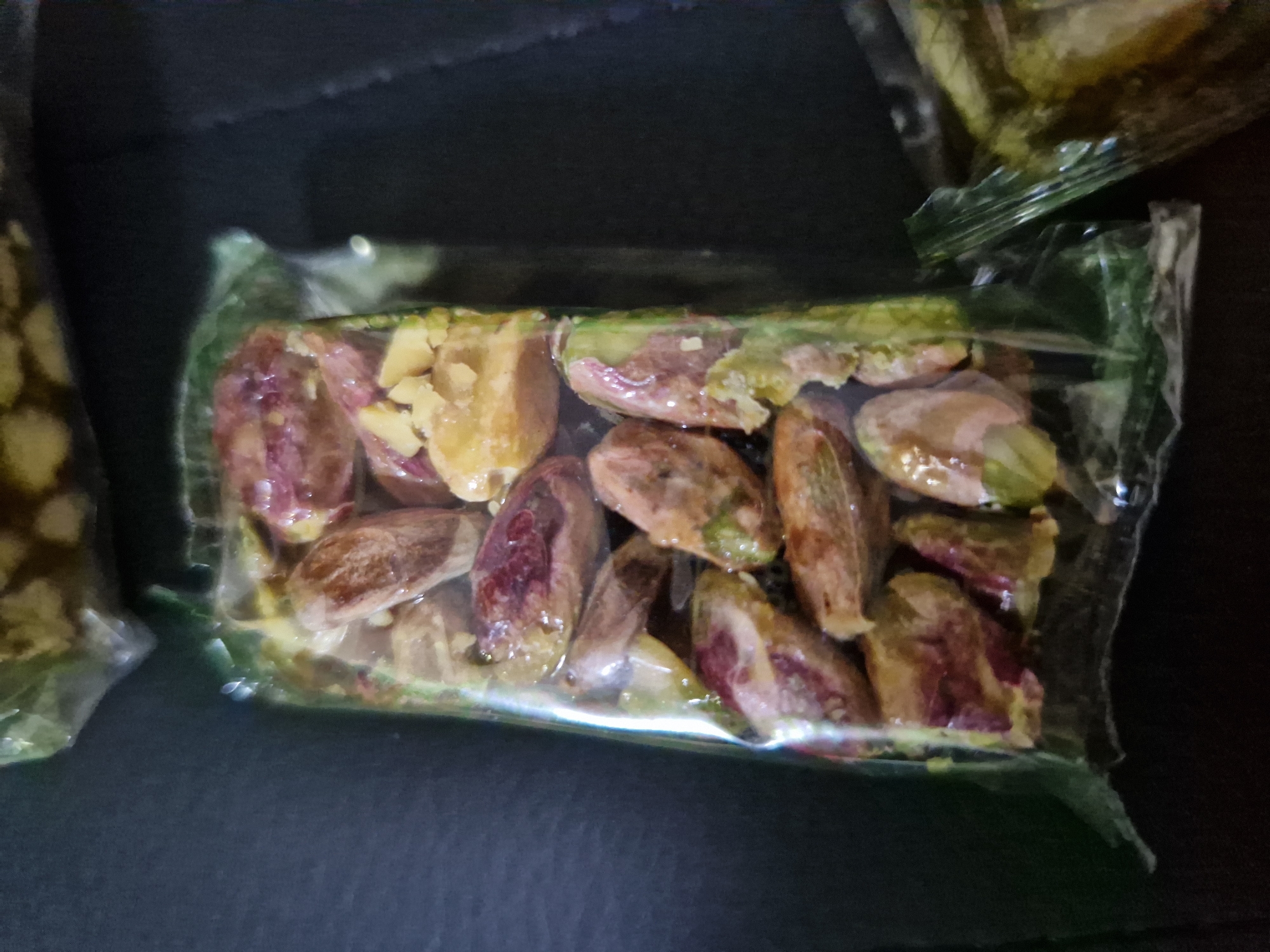 The image displays a close-up, overhead view of several packages of pistachios. The pistachios are shelled and appear to be slightly oily, with visible purple and green hues. They are packed tightly within transparent plastic packaging. One package is centrally located and appears to be a rectangular bar shape, filled with the nuts. Another partially visible package is to the right, and a third is at the top. The background is dark and out of focus, suggesting a simple surface. There are no people or actions depicted, and the scene is static. The provided location, Sofia, Bulgaria, does not correspond to any visual cues within the image.