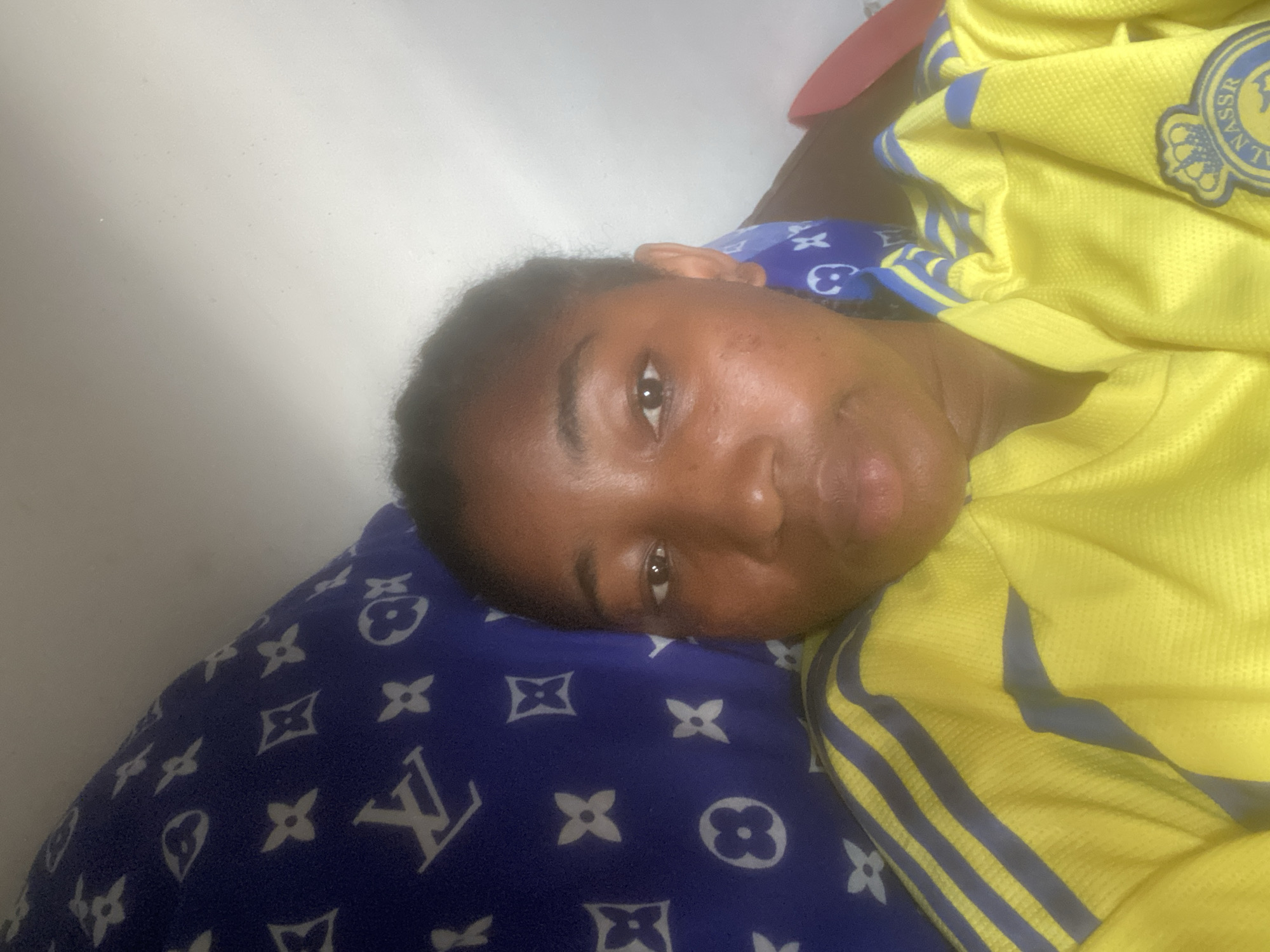 A close-up indoor shot depicts a person resting on a blue pillow patterned with white "LV" logos, possibly in a domestic setting. The person is partially covered by a yellow jersey with blue stripes and an emblem on the right side, and the background features a white wall. The location is unknown and not possible to determine from the image, though it was requested to be in Lokoja, Nigeria. There is no forensic information available.