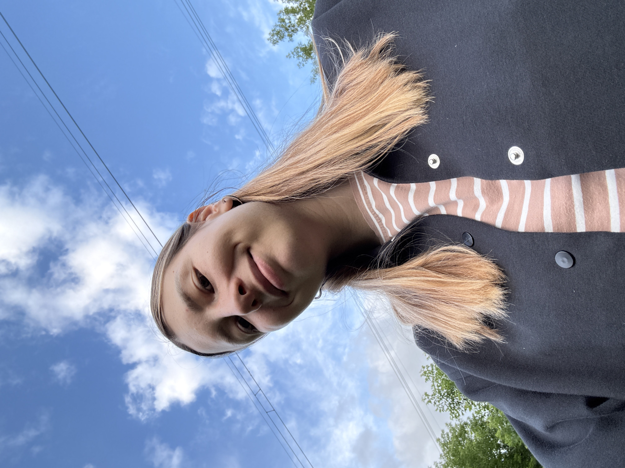 A low-angle, medium shot captures a young Caucasian woman taking a selfie outdoors on a cloudy day. She has light brown hair styled down, and is wearing a dark jacket over a striped shirt. In the background, trees and power lines are visible against a blue sky with white clouds. The setting appears to be a park or residential area, likely within Moscow, Russia, given the presence of power lines and deciduous trees common to the region.