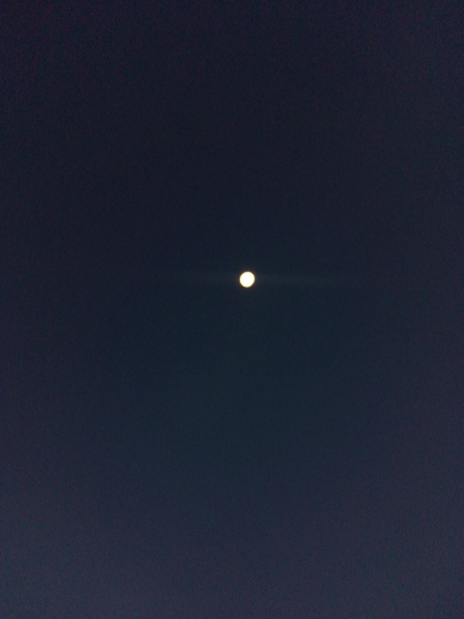 The image displays a clear night sky over Juan Griego, Venezuela. A single, prominent, bright circular celestial body, identified as the moon, is centrally positioned within the dark, largely uniform expanse. A subtle horizontal light streak emanates from the moon, extending slightly to the right. No other celestial bodies, terrestrial features, artificial structures, individuals, or discernible activities are present.