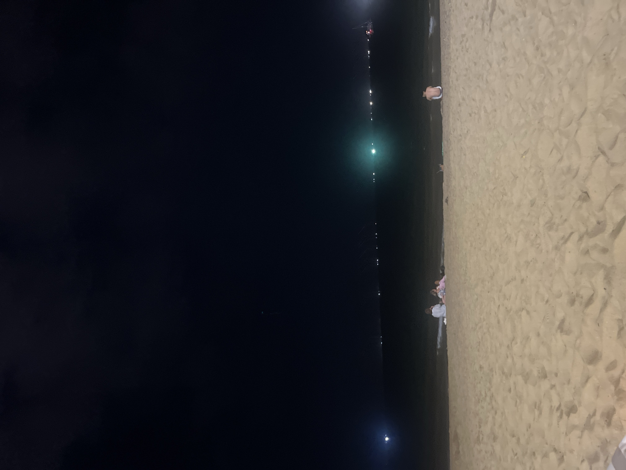 The image depicts a nighttime scene on a sandy beach in Nha Trang Ward, Vietnam. The sky is dark, indicating it is nighttime. The water of the sea is visible, with some gentle waves reaching the shore. In the foreground, the sandy beach is illuminated by an unseen light source, casting a soft glow on the sand.
Several individuals are present on the beach. In the middle ground, a group of three people are seated on the sand, facing the sea. To their right, further down the beach, another person is sitting facing away from the camera, towards the water. In the distance, across the sea, a line of lights suggests a developed area or harbor. There are also some lights on the water that could be from boats. The overall atmosphere is quiet and serene, with the sound of the waves being the only prominent element.