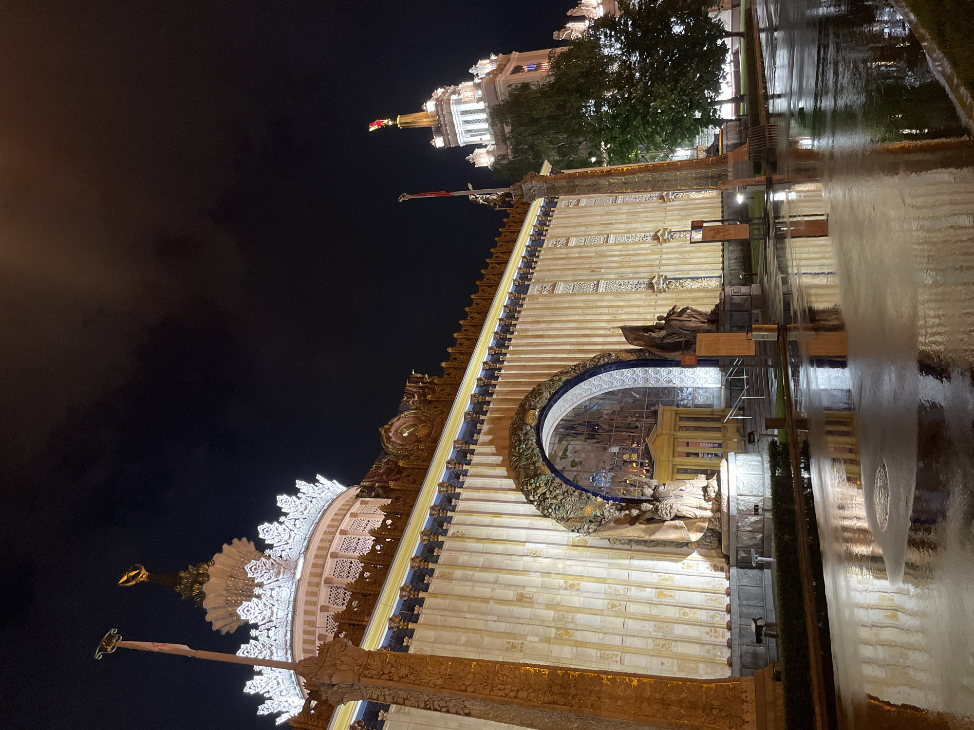 The image shows a building at night, likely located in Moscow, Russia, based on the architectural style. The building features ornate white facades with gold detailing and decorative elements. There's a large, rounded archway with intricate carvings and sculptures. The lower part of the scene includes a reflective surface, possibly a wet pavement, creating a mirror-like effect of the building. In the background, there's another illuminated building with a tower and a red flag on top. The sky is dark, suggesting it's nighttime.