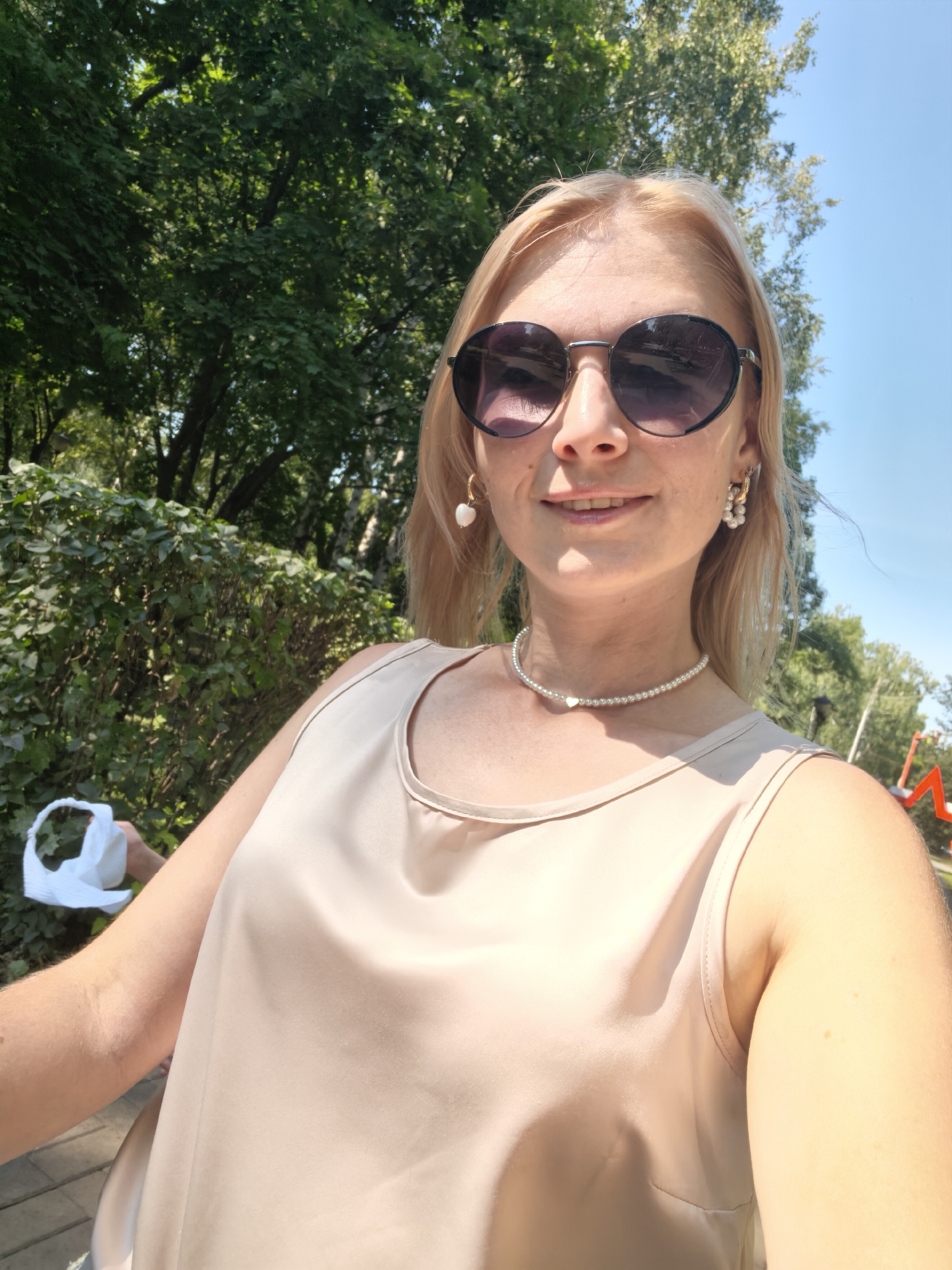 A young woman with blonde hair and sunglasses takes a selfie outdoors. She is wearing a beige sleeveless top, a pearl necklace, and pearl earrings. The background consists of green trees and a blue sky. The image is taken in a park-like setting with a paved path visible in the foreground. There is a red structure with a star visible in the far right background, which may be part of a playground or park decoration. The woman is holding a white object, possibly a bag or a piece of clothing, in her left hand.