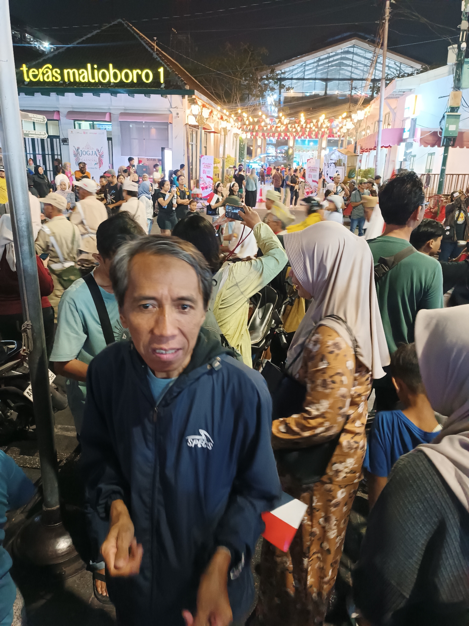 A nighttime outdoor street scene in the Malioboro 1 area of Yogyakarta, Indonesia, features a crowd of people gathered under illuminated string lights and building signage. A prominent sign reads "teras malioboro 1" in yellow lettering. People of various ages and genders are present, some walking, some standing, and some interacting. In the foreground, an elderly man in a dark blue jacket is looking at the camera, with his hands held in front of him. Behind him, a woman in a tan hijab and floral dress is visible. Other individuals are seen in the background, some holding phones, suggesting they might be taking pictures or videos. The overall atmosphere appears to be lively, possibly during an event or festival, given the decorations and the density of people. The environment suggests a commercial or entertainment district, characteristic of Malioboro.