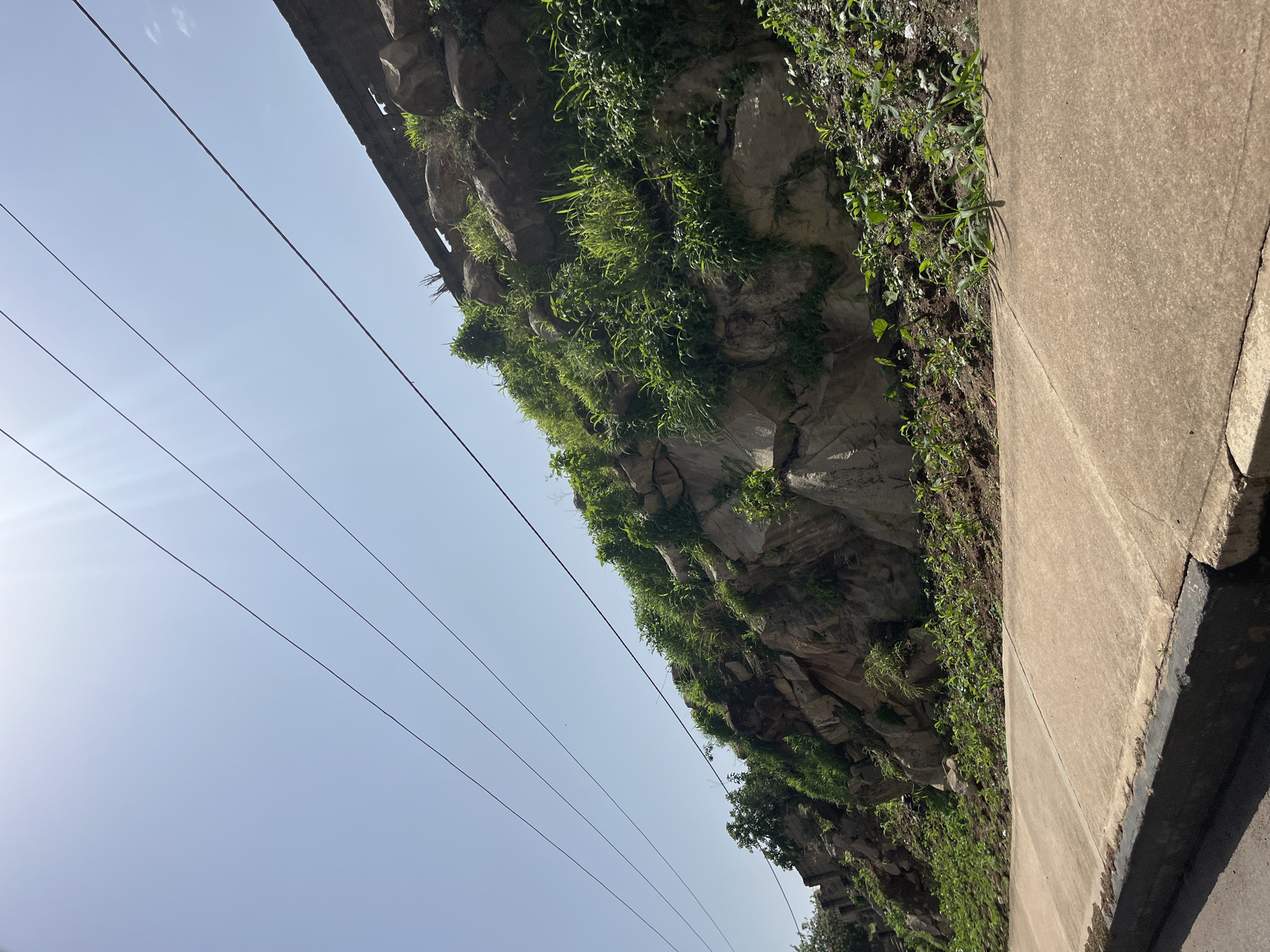 A low-angle outdoor shot captures a rocky hillside with sparse vegetation, possibly a natural or man-made embankment, adjacent to a paved sidewalk. Four parallel power lines stretch across the bright, clear sky. The rock face is irregular and layered, with patches of green grass and small plants growing in crevices and at the base. The sidewalk, made of concrete, shows some wear and tear, with a dark-colored curb separating it from a darker paved surface, possibly a road, in the foreground. The lighting suggests a sunny day. The location is identified as Bukuru, Nigeria. There are no people visible in the frame, and no actions or interactions are taking place.