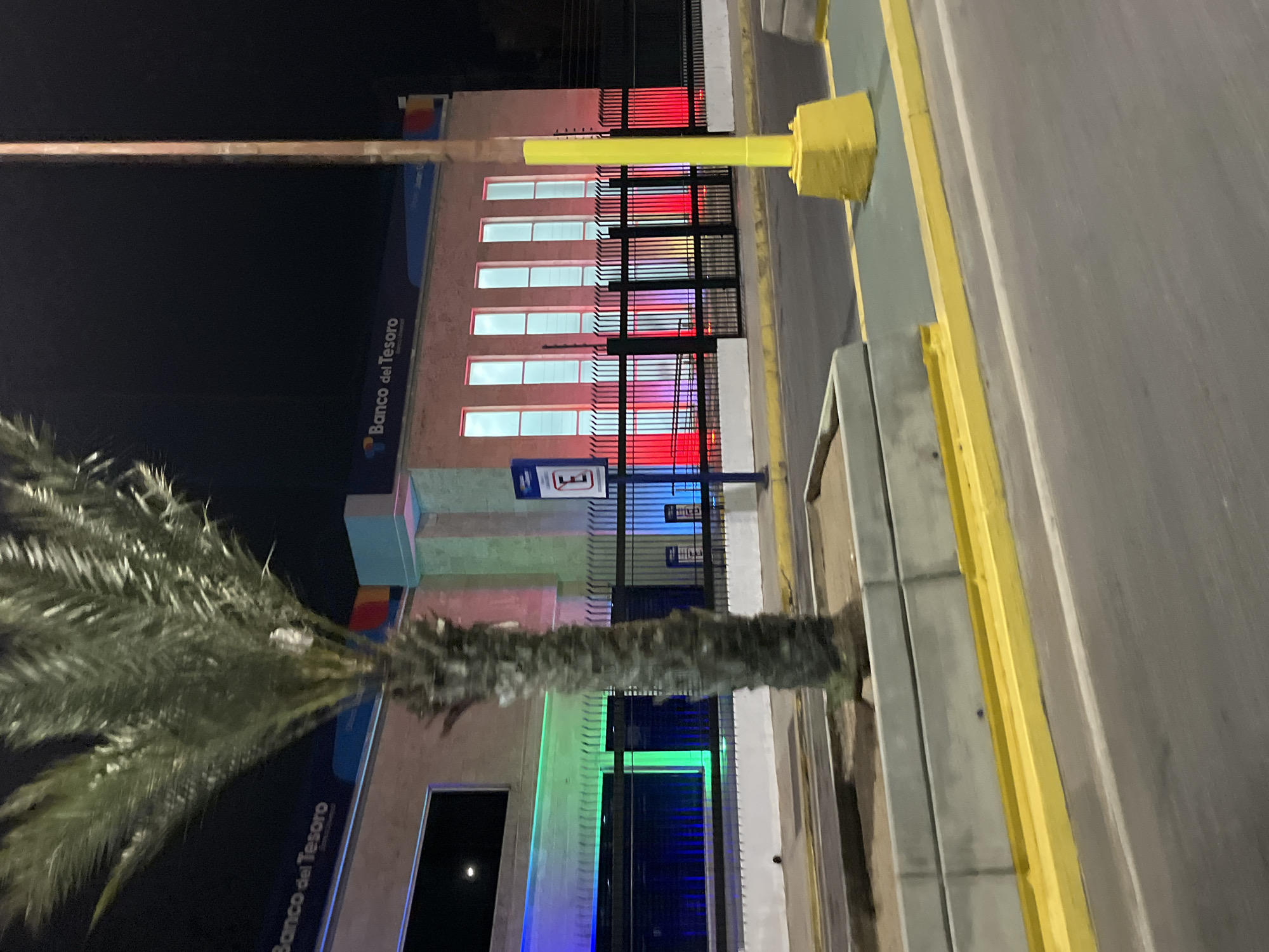 At night, the Banco del Tesoro branch in Juan Griego, Venezuela, displays illuminated, colorful lights that illuminate the building. A tall palm tree stands near the building, its leaves obscuring some parts of the facade. A sidewalk with a yellow curb runs along the side of the building.