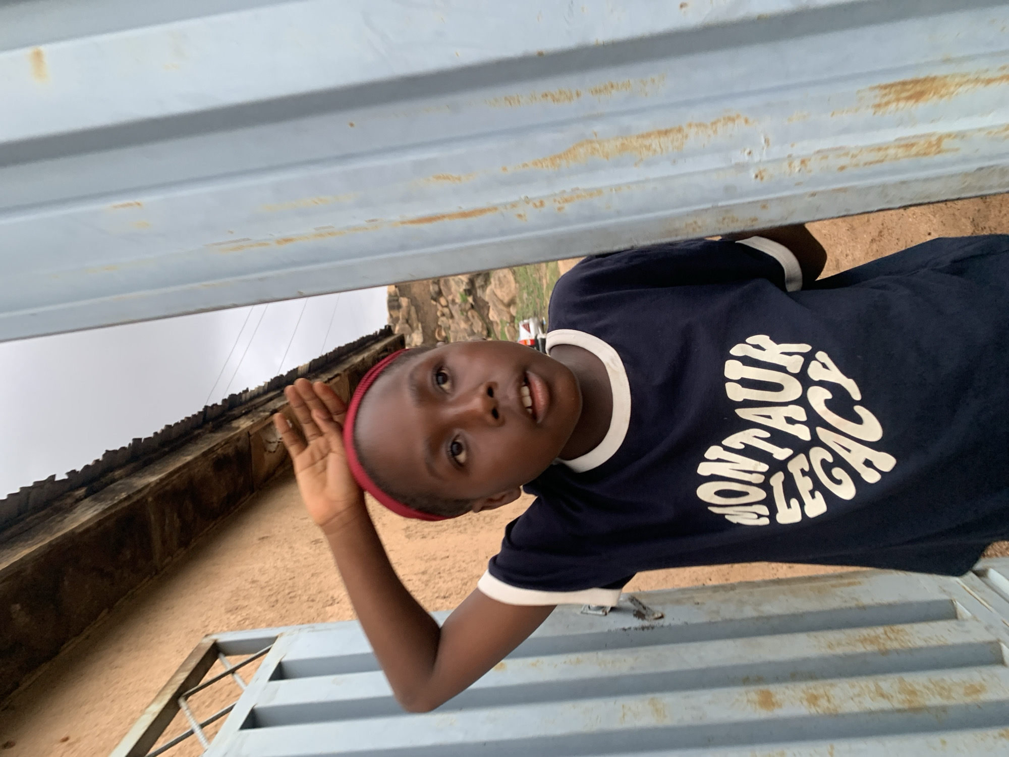 A young person is captured in a candid photograph, partially obscured by a light blue metal fence. The individual has dark skin, a red headband, and is wearing a navy blue t-shirt with the phrase "MONTAUK LEGACY" printed on the front in white lettering. They are looking directly at the camera with a slightly open mouth. Their right arm is raised with their hand facing outwards. The background includes a glimpse of a building with a weathered roof, and a natural landscape with rocks and vegetation. The location is Jos, Nigeria.