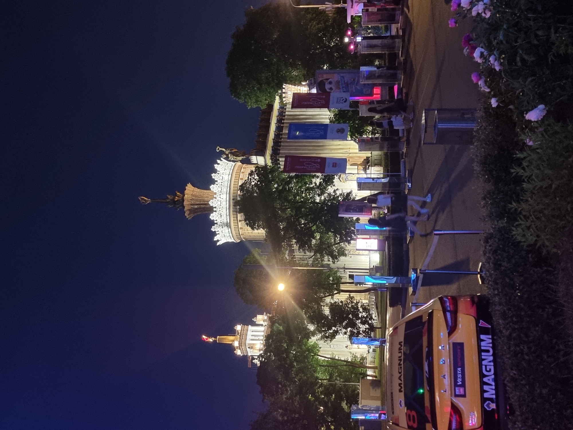 The image depicts an outdoor scene at night, likely at the VDNKh (Exhibition of Achievements of National Economy) in Moscow, Russia, given the architecture. In the foreground, a yellow racing car with "MAGNUM" branding on the rear is parked. Behind the car, several people are walking in the middle ground, illuminated by various light sources. Tall, vertical display stands with various graphics are positioned in the area. In the background, impressive illuminated buildings with ornate architectural features, characteristic of Soviet-era exhibition pavilions, are visible against a dark sky. Trees are scattered throughout the scene, some of which are lit by the artificial lights. The overall atmosphere is one of a public event or exhibition taking place after dark.