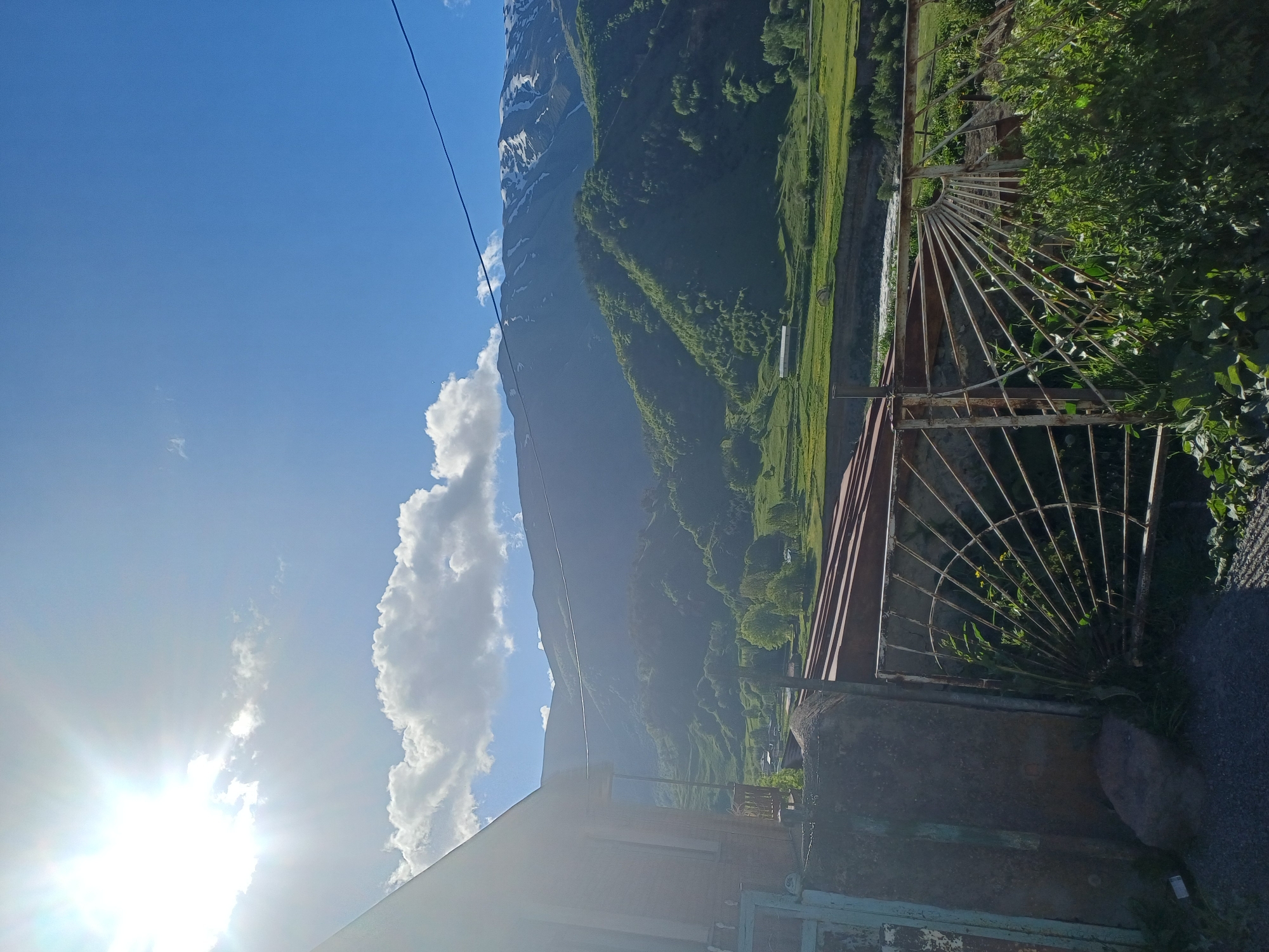 The image depicts a daytime outdoor scene with a bright sun and blue sky. In the foreground, a portion of a building with brown siding and a section of a rusted metal gate are visible on the left. The gate features a decorative, fan-like design made of metal bars. Beyond the gate, lush green vegetation and a river are visible. In the background, rolling green hills rise to meet a large mountain range, the peaks of which are capped with snow. The overall impression is of a rural or mountainous landscape, possibly in a location like Goristsikhe, Georgia, given the terrain and architecture. There are no people or animals visible in the image.