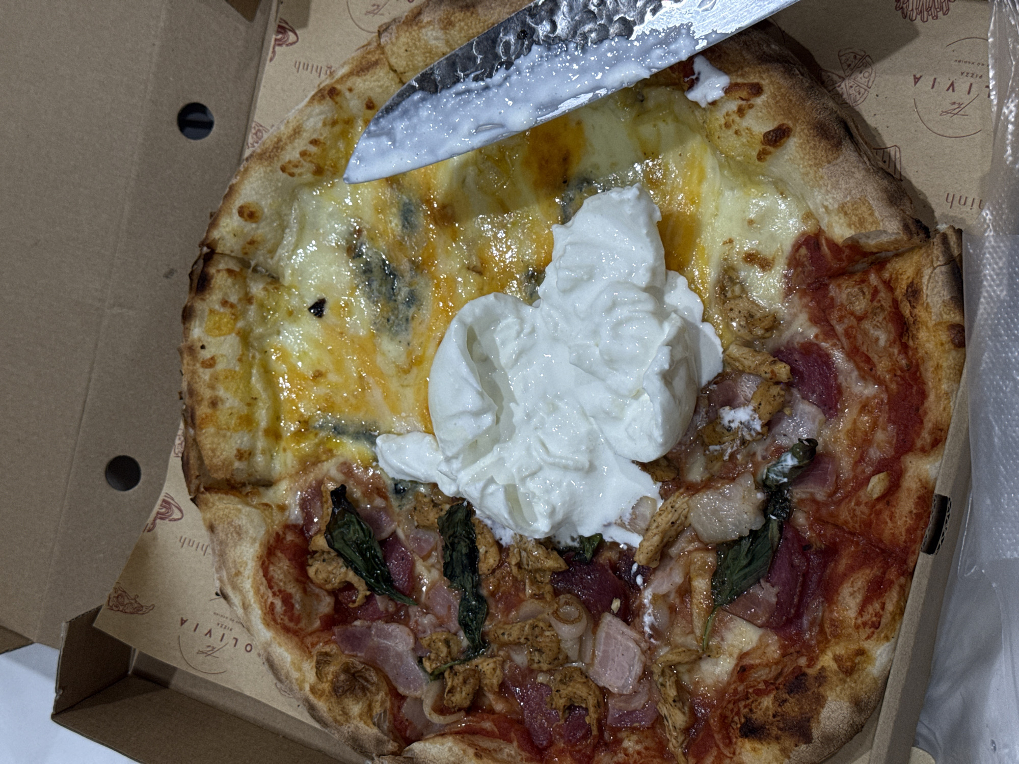 A pizza in a cardboard delivery box is being cut with a silver knife. The pizza is topped with cheese, sauce, meat, and a mound of white cream cheese. The delivery box has the logo of "Olivia Pizza" and the text "Pizza ao Heahin" and "Đà Nẵng, Vietnam" printed on it.