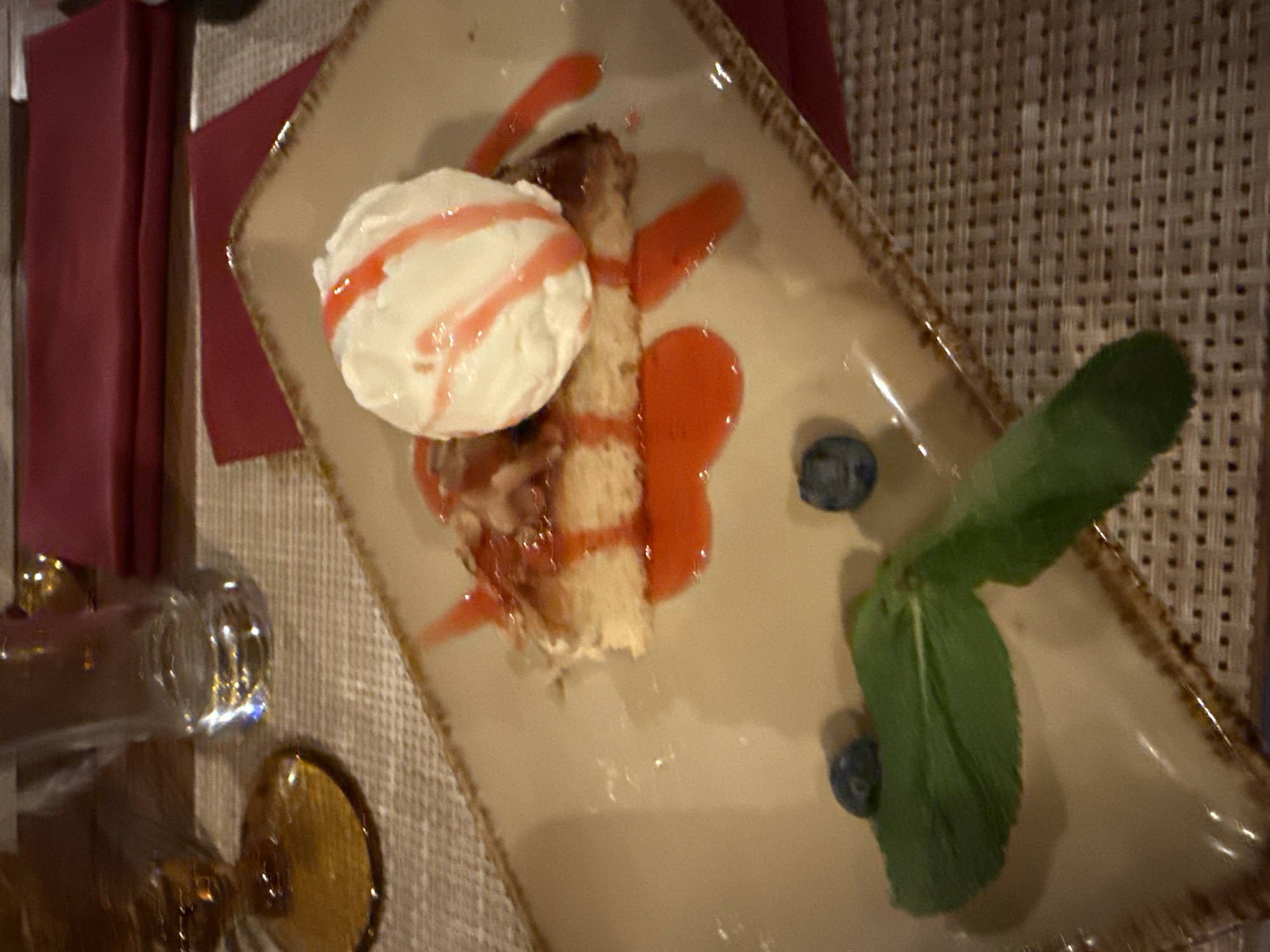 The image shows a plate of dessert sitting on a woven surface, possibly a placemat. The dessert consists of a slice of cake, topped with a scoop of white cream with red sauce drizzled over it, alongside red sauce drizzles on the plate. Two blueberries and a sprig of green leaves garnish the plate. To the left of the plate, there is a glass and a golden-colored object. The setting appears to be indoors, possibly a restaurant. There is no information that allows me to determine the city in which this image was taken, and I cannot confirm the location.