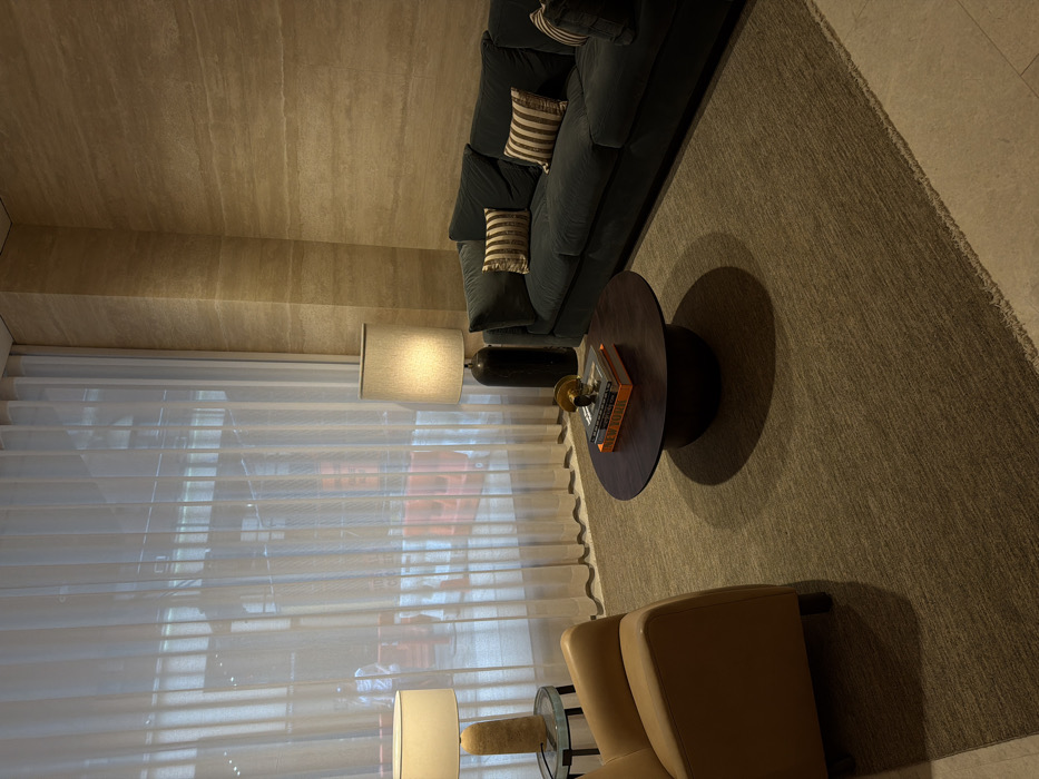The image depicts an interior lounge or waiting area within a building in New York City, United States of America, looking out onto an urban street scene.
The interior is furnished with a dark teal velvet sofa positioned against a textured light brown stone or travertine wall, and a tan leather armchair. A round, dark brown wooden coffee table sits on a dark beige textured area rug in the center of the room. On the coffee table, a stack of books, including one with "NEW YORK" visible on its spine, is topped by a small, abstract golden sculpture. Two illuminated table lamps are present: one with a light-colored, textured base and white shade on a small, round glass-topped side table next to the armchair, and another with a tall, dark cylindrical base and light grey shade standing on the floor between the sofa and the window.
A large window, spanning the left side of the room, is covered by sheer white floor-to-ceiling curtains. Through these translucent curtains, an exterior street scene is partially visible. Multiple orange construction barriers are lined along a street or sidewalk. A single, indistinct person wearing a light-colored shirt is visible standing near these barriers. No specific actions or interactions are occurring within the immediate interior space.