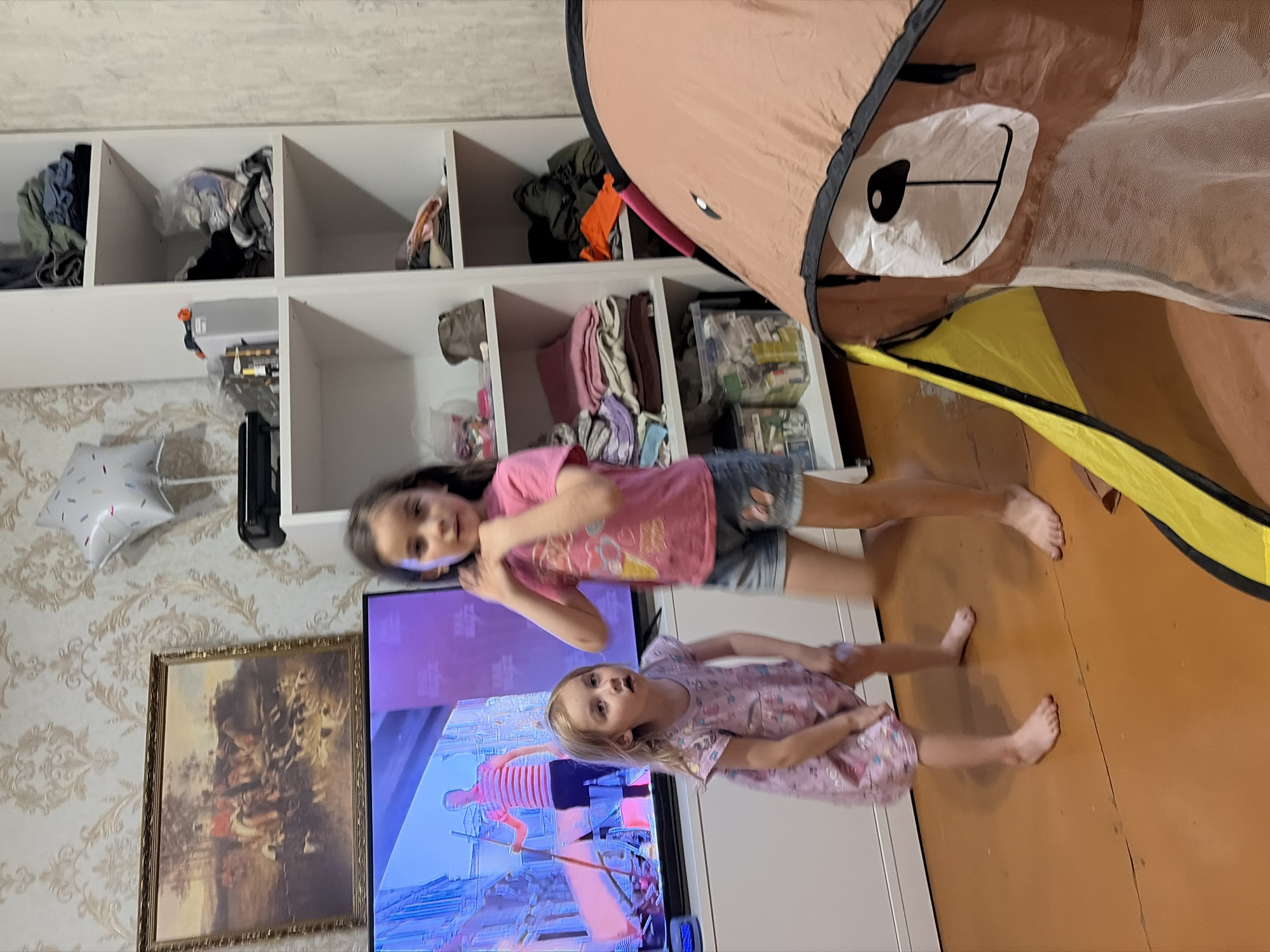 The image depicts an indoor scene featuring two young girls in a room with a television, a bookshelf, and a play tent. The younger girl, on the left, wears a pink floral dress and stands barefoot with her legs slightly bent. The older girl, on the right, is wearing a pink t-shirt and denim shorts and is captured mid-movement, with one arm raised.
The room appears to be a living area, possibly a child's playroom. The television screen shows a colorful graphic with a person on it. To the left of the television, a framed painting of a hunting scene hangs on a wall with patterned wallpaper. A white bookshelf unit filled with clothes and various items is visible to the right. In the foreground, a pop-up play tent with a bear face design is partially visible. The floor is a warm-toned, possibly wooden surface with some visible cracks.
There are no specific elements in the image that would allow for the identification of the city of Tambov, Russia, or any other specific geographic location.