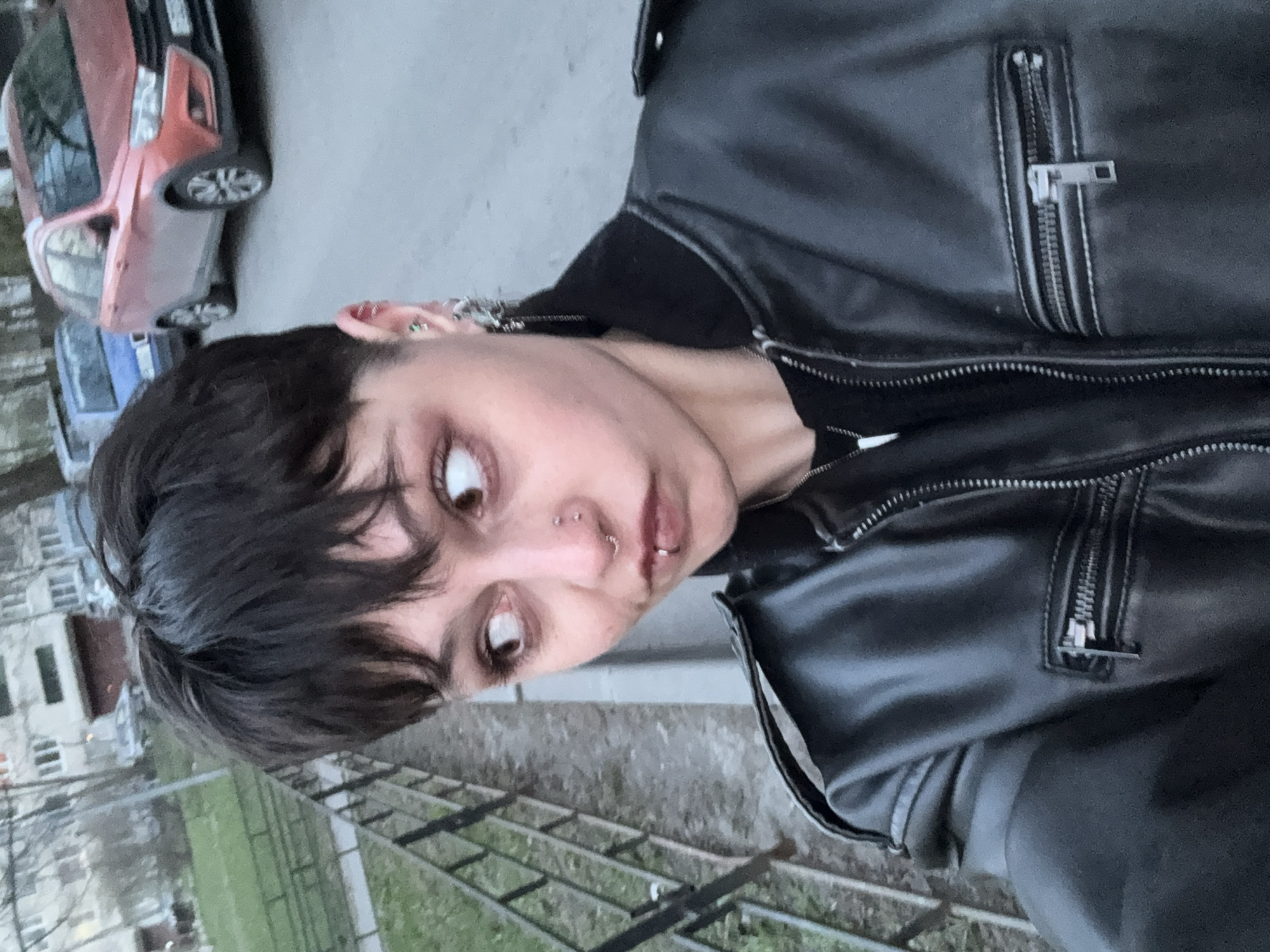 A person with short, dark hair is depicted in an outdoor setting. They are wearing a black leather jacket and have multiple piercings on their ear, nose, and lip. Buildings and vehicles, including a red SUV and a blue car, are visible in the background. Based on the architectural style and the presence of typical urban elements, the location is suspected to be in the city of Saint Petersburg, Russia. The photo is taken from a low angle, causing the person to appear to be looking down at the camera.