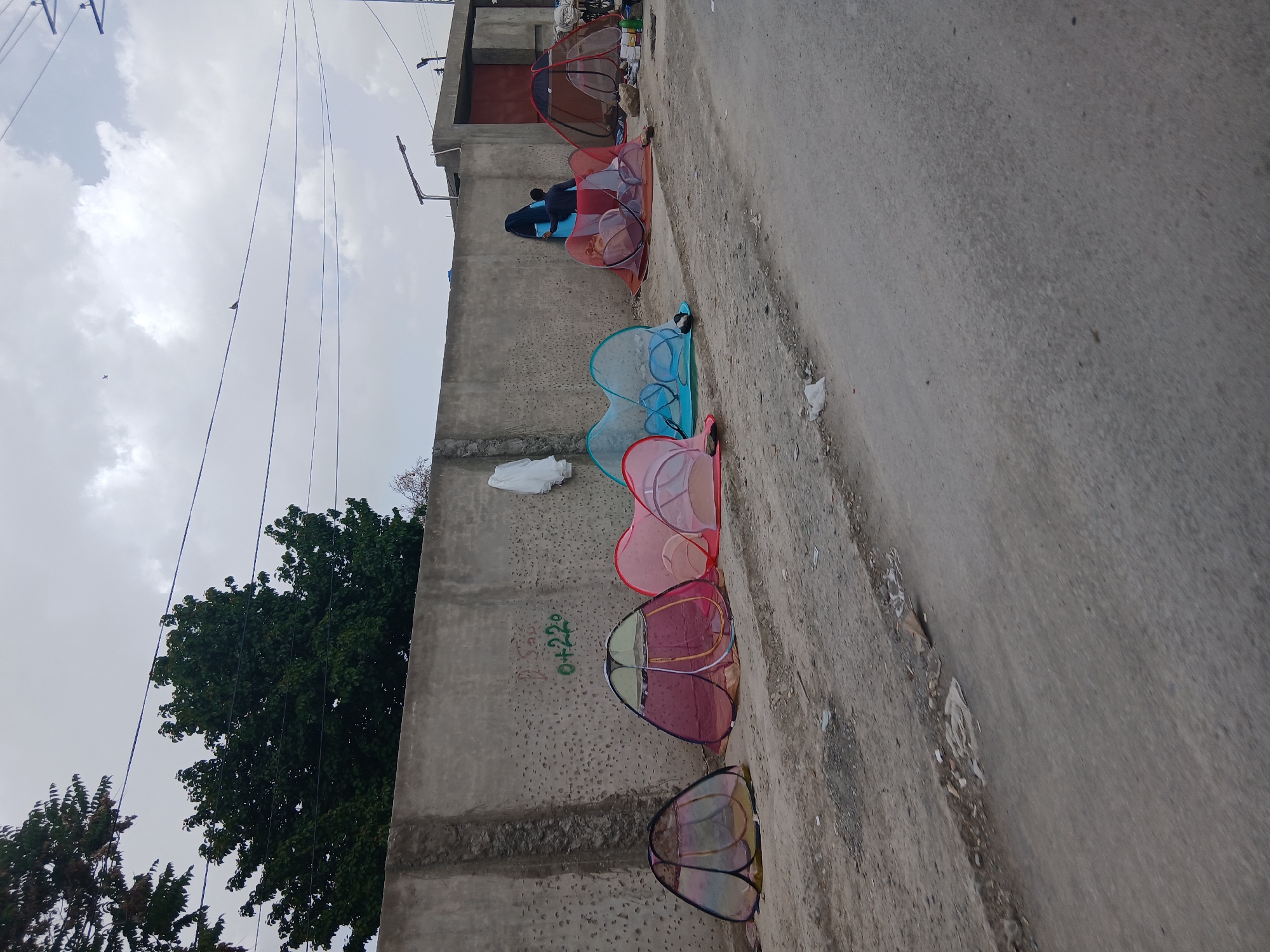 The image, taken in Quetta City Tehsil, Pakistan, shows a series of small, collapsible, colorful shelters or tents lined up against a concrete wall. The tents appear to be made of netting or mesh material, with different color combinations of red, blue, pink and other colors. A person is visible near the tents at the far end. The wall appears to be a boundary or retaining wall alongside a paved street or road. Green trees are visible behind the wall on the left side. The sky is overcast.