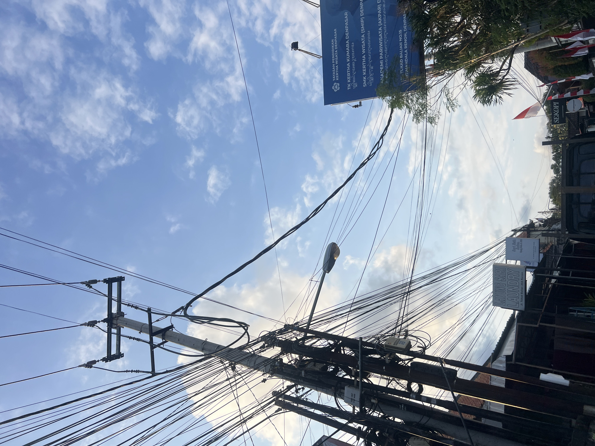 The image captures a view of a street in Denpasar, Indonesia, dominated by a complex network of overhead power lines and a utility pole. The sky above is a bright blue with scattered white clouds, suggesting it is daytime. A street lamp is attached to the utility pole. On the right side of the image, a large blue sign is visible, featuring text in both Indonesian and what appears to be Balinese script, listing educational institutions like "TK KERTHA KUMARA DENPASAR" and "SMK KERTHA WISATA (SMIP) DENPASAR". Further down, the sign indicates an address as "JL. TUKAD BALIAN NO.15". Below this sign, several Indonesian flags are displayed. On the left side, a sign for "LAYUNG SARI LAUNDRY" with an address "JL. Tukad Balian 20A Renon" is visible, alongside other shop fronts. Trees and foliage are also present, with the architecture suggesting a typical urban environment in Indonesia. The overall impression is one of a busy street scene, characteristic of a bustling city in Bali.