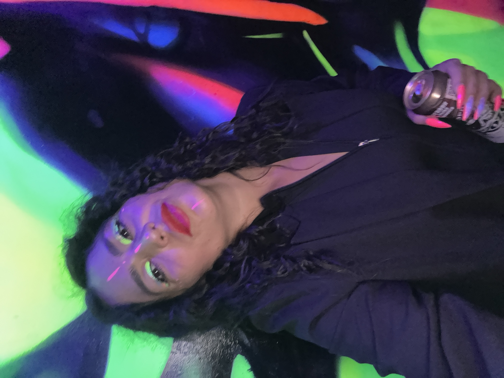 A woman lies against a backdrop of brightly colored graffiti art. The art features neon green, orange, blue, and black, with some areas appearing to glow. The woman is fair-skinned with dark, curly hair and red lipstick. She has a stripe of pink paint on her forehead and under her eyes. She is wearing a dark-colored zip-up jacket or shirt. Her nails are long and pointed, with a neon pink color that appears to glow under the lighting. She is holding a can of beverage in her right hand, which also shows the effects of the bright, colorful lighting. The image has a strong lighting effect from the graffiti, casting different hues on the woman's face and clothing. The location is Chaikovsky, Russia.