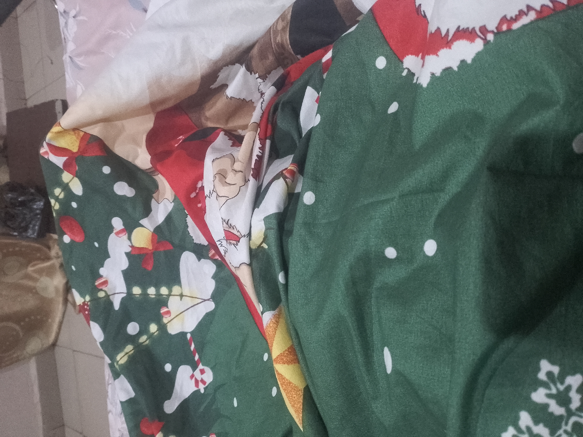 A close-up, overhead shot captures a pile of fabric with a Christmas theme. The fabric is folded and wrinkled, obscuring a clear view of any full objects. The dominant colors are deep green and red, with white accents. On the green fabric, there is a repeating pattern of white dots, reminiscent of snow or tinsel. Interspersed with the dots are stylized white figures that appear to be snowmen or possibly Santa Claus figures. Red bows and yellow elements are also visible within the pattern. A portion of red fabric with a white, jagged edge, suggesting a Santa hat or coat trim, is visible in the upper right quadrant. The overall impression is a festive, possibly bedding or drapery material, with a holiday design. The image is taken in an indoor setting, likely a home, with a muted background.