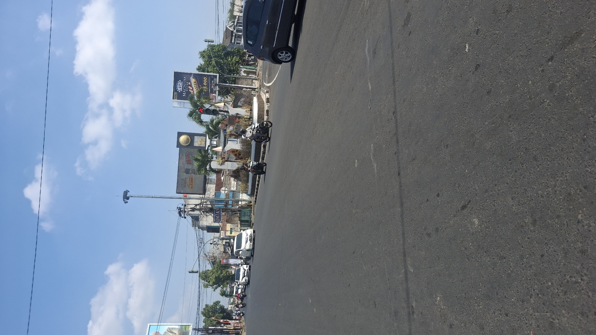The image shows a street intersection in Tasikmalaya, Indonesia, on a sunny day with a clear blue sky and scattered white clouds. Several vehicles are present, including white cars, a dark gray car, and motorcycles. Pedestrians are not clearly visible. Buildings and advertisements are on the sides of the road, including billboards with text in Indonesian, one of which advertises "MAGNUM FILTER" for "Rp. 5,000". There is a traffic light that appears to be red. The road surface is asphalt, with visible tire marks and some debris. The general environment suggests a moderately busy urban area.