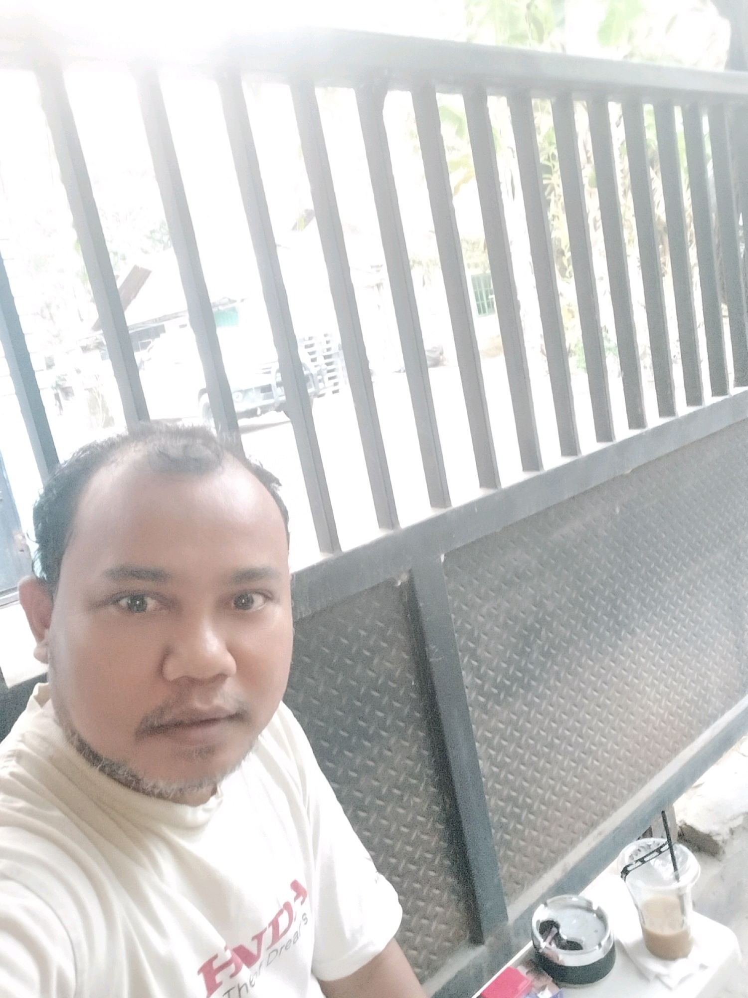 A man with a receding hairline and a light stubble around his chin and mouth is taking a selfie outdoors. He is wearing a white t-shirt with red lettering. In the background, a metal fence with vertical bars runs across the frame, with foliage and a building visible through the gaps. To the right of the man, on a table, are a drink in a clear plastic cup with a straw and an ashtray with a cigarette butt. The scene appears to be in a public outdoor area.