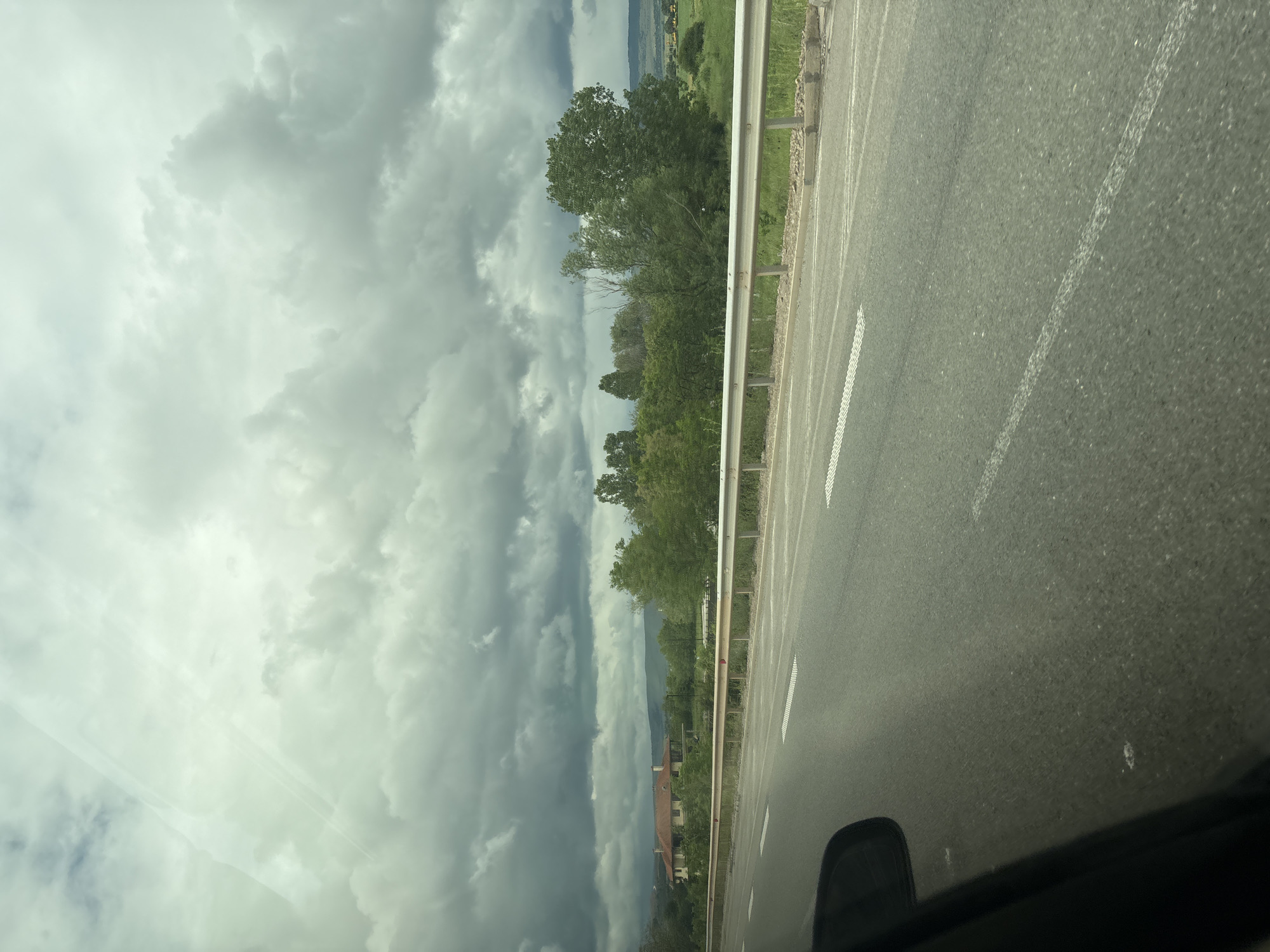 The image, viewed as if correctly oriented, captures a landscape scene from the perspective of a moving vehicle. An asphalt road, featuring a white dashed line for lane separation and a solid white line on the shoulder, dominates the right side and foreground. A metal guardrail runs parallel to the road, bordering a green, undulating landscape.
To the left of the road and guardrail, numerous tall, leafy trees with vibrant green foliage cover the middle ground. Beyond these trees, the terrain extends into distant hills or mountains. Scattered across the landscape in the mid-distance are buildings with reddish-brown tiled roofs, suggesting a rural settlement or village, consistent with the location context of Hrabarsko, Bulgaria.
The sky is largely filled with heavy, dark grey, overcast clouds, indicative of pre-storm or stormy weather. Diffused light filters through lighter patches in the cloud cover, and subtle reflections, likely from a car's windshield, are visible in the upper left portion of the sky. The bottom right corner of the frame shows a dark section of the car's interior, such as a dashboard or door panel.
The scene depicts a daytime journey through a verdant, hilly countryside under a dramatic, cloudy sky. No text is visible in the image.