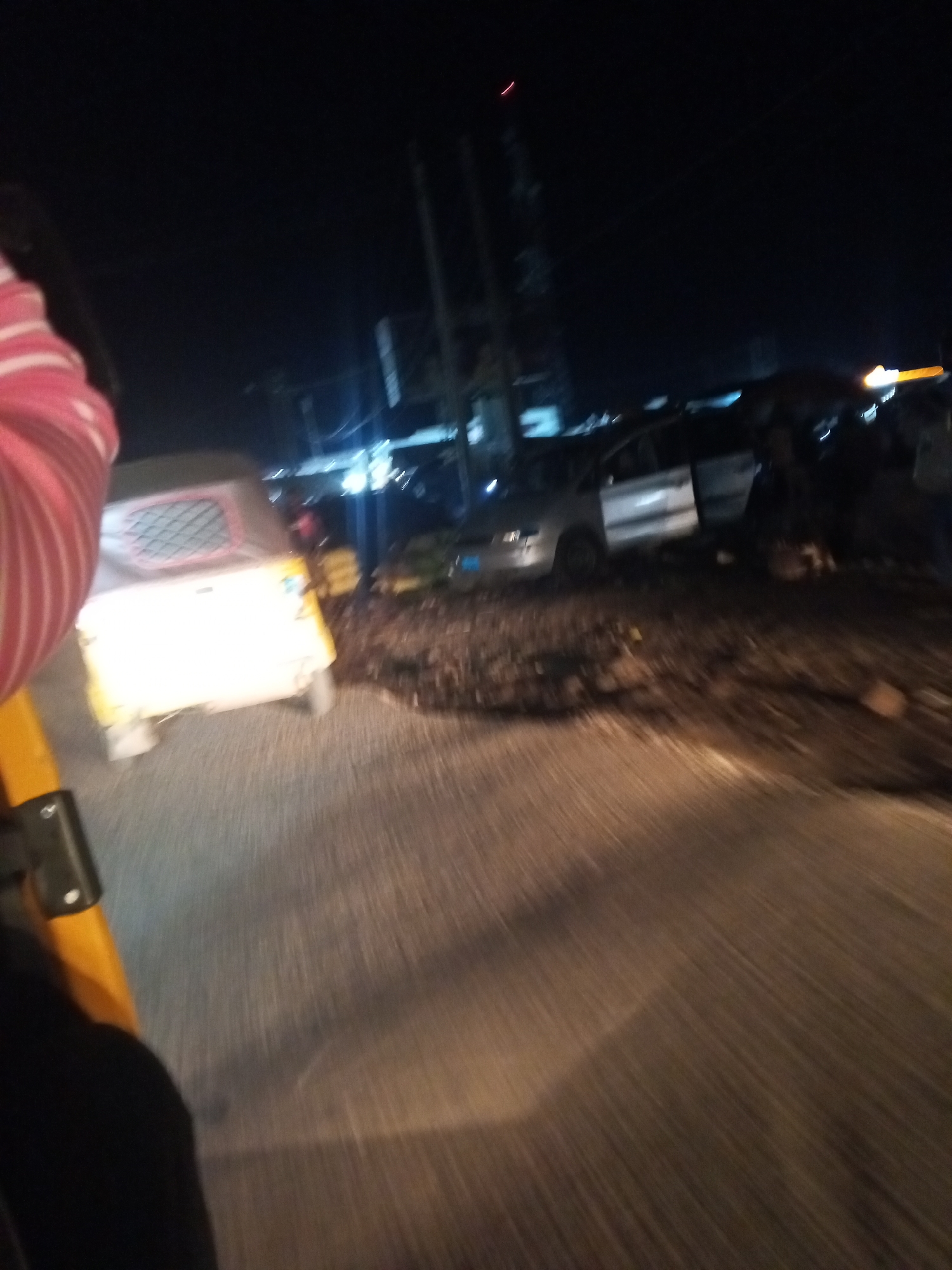 At night in Lokoja, Nigeria, an auto-rickshaw is parked on the side of a road. The auto-rickshaw is yellow and white with a red-trimmed window. The area around the auto-rickshaw is uneven and appears to be a mixture of dirt and debris. In the background, a silver car is parked with its doors open. There are lights visible in the distance, suggesting the presence of other vehicles or structures. The scene is dimly lit, with some parts of the image appearing blurry due to motion or low light conditions.