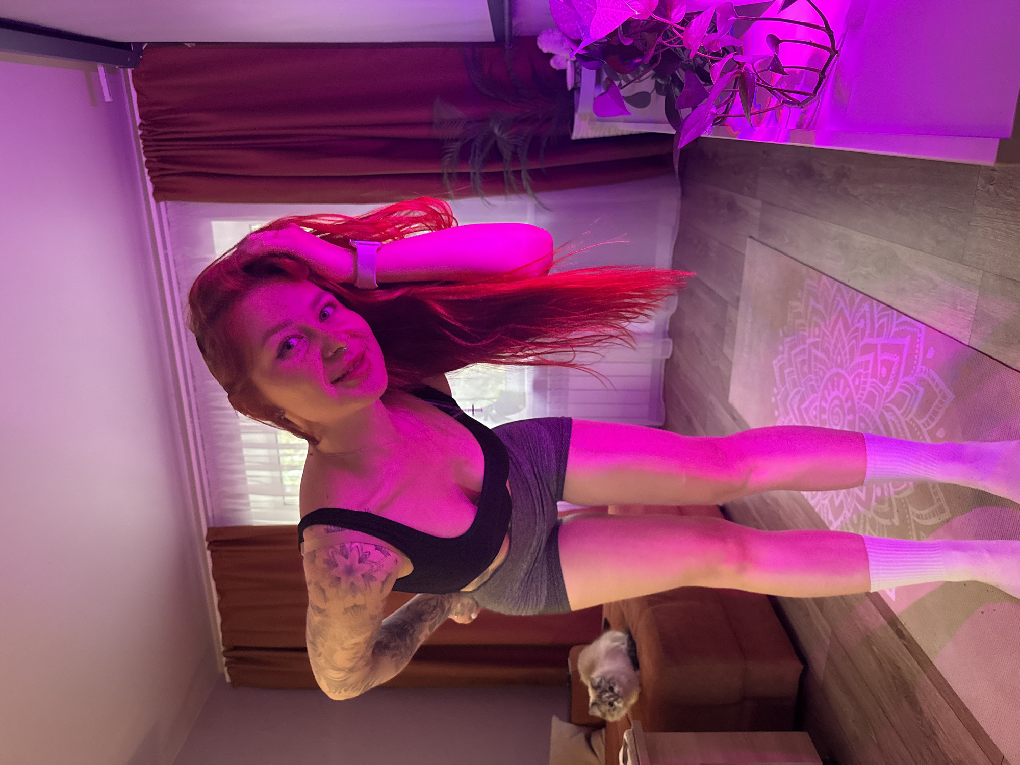 A woman with red hair and tattoos on her arm is standing indoors in what appears to be a room with a yoga mat on the floor. The room is lit with a purple light, casting a colorful hue over everything. The woman is wearing a black sports bra and grey shorts, and white socks. The background includes a window, curtains, and a plant on a shelf. There is also a cat visible in the lower part of the image. There's no specific evidence to place the location within Rostov-on-Don, Russia.