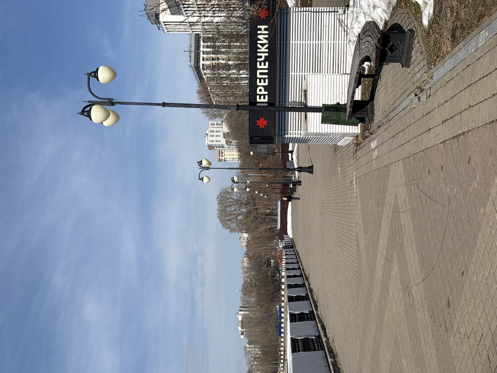The image shows a street view in Izhevsk, Russia. The photograph is taken from the sidewalk, looking towards a street lined with bare trees and buildings in the background. A row of black lampposts with egg-shaped lights is visible along the sidewalk. A sign with Cyrillic lettering, reading "ПЕРЕПЕЧКИН" is displayed perpendicular to the sidewalk. A bench is placed adjacent to the sidewalk. The sky is blue with scattered clouds. The sidewalk is paved with patterned bricks. There is snow next to the sidewalk in certain areas.