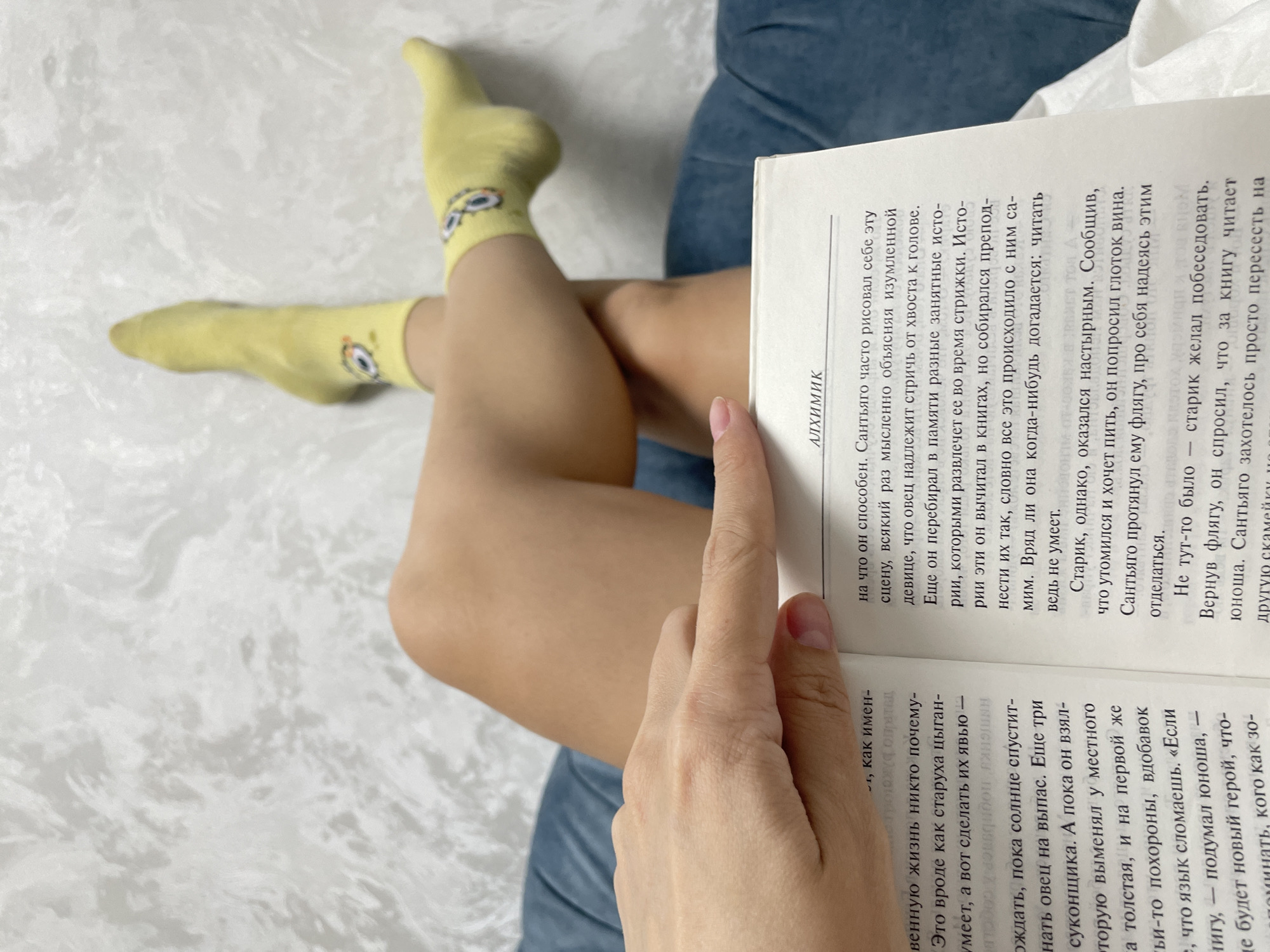 A person is sitting with crossed legs, wearing yellow socks with cartoon eyes, and reading a book in Russian. The book is titled "Алхимик" (The Alchemist). The person's hand is holding the book open, and the text on the page is visible. The background is a neutral color, possibly a bed or couch. The location is unknown, but the text indicates it is in Russian, which could possibly be in the city of Izhevsk, Russia.