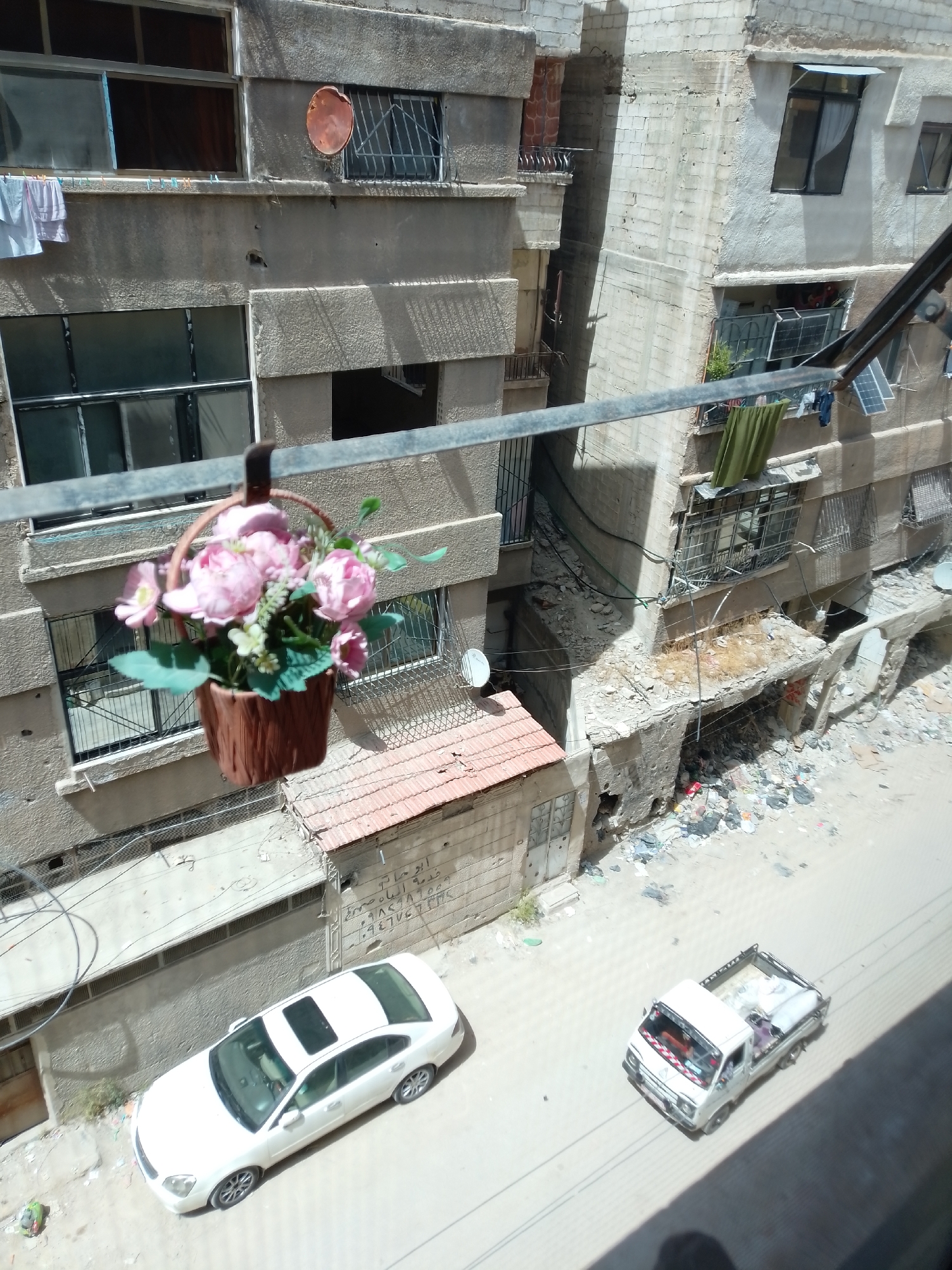 The image shows an aerial view of a street in Zamalka, Syria. The foreground features a hanging basket of artificial pink flowers. Behind the flowers, multi-story buildings show signs of damage and disrepair. Some buildings have satellite dishes mounted on their facades. Debris and rubble are visible at the base of some buildings. Two vehicles are parked on the street: a white sedan with a sunroof and a small white truck. There are no people visible in the image. The environment appears to be urban, and possibly post-conflict, as evidenced by the building damage.