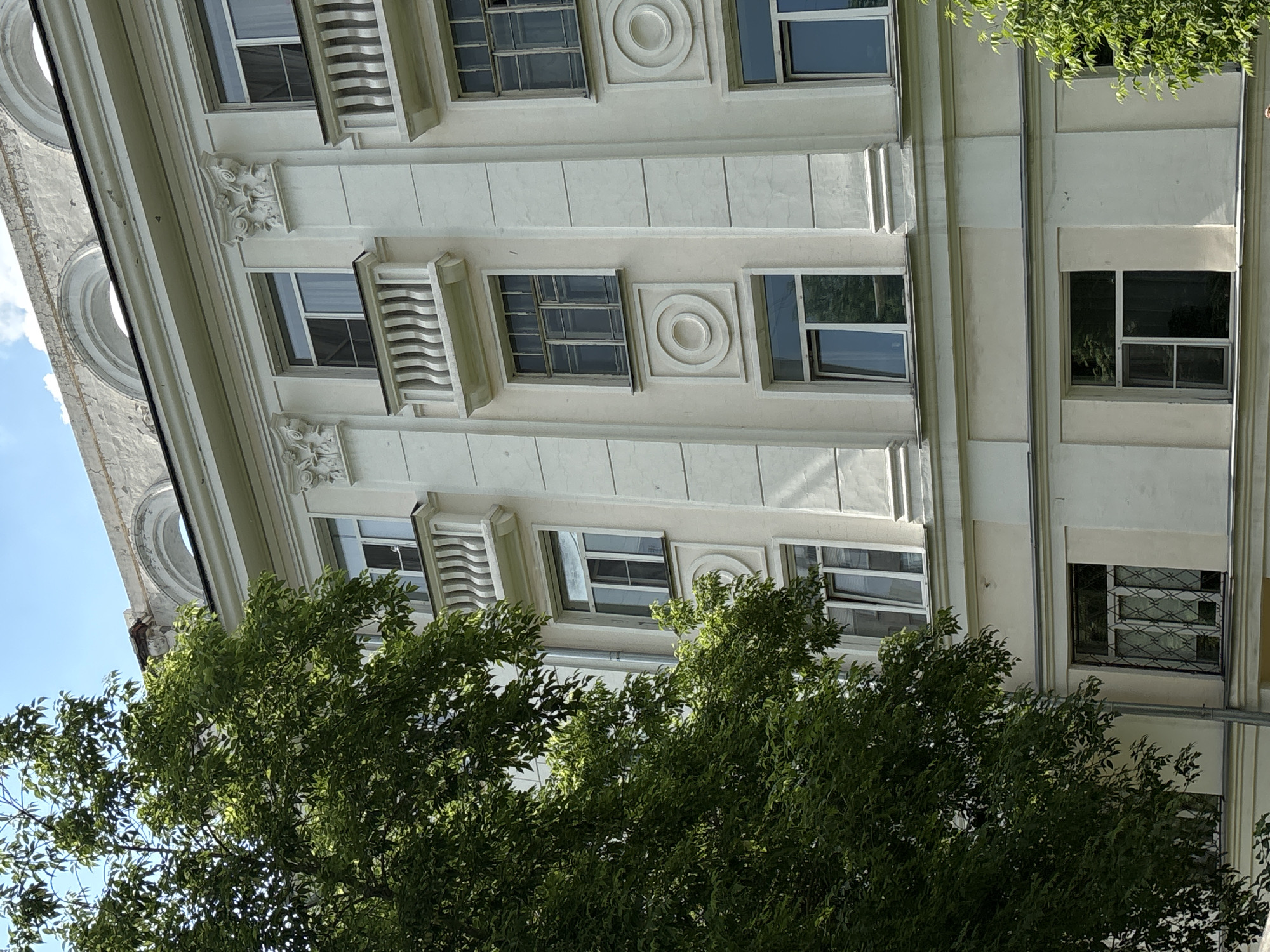 The image shows the facade of a light-colored, multi-story building in Novosibirsk, Russia. The building features neoclassical architectural elements, including decorative columns, cornices, and arched window frames. Several windows are visible, some with balconies and ornate railings. Lush green trees partially obscure the left side of the building and also appear on the right. The sky above is bright blue with a few white clouds. The image is taken from a low angle, looking upwards at the building.
