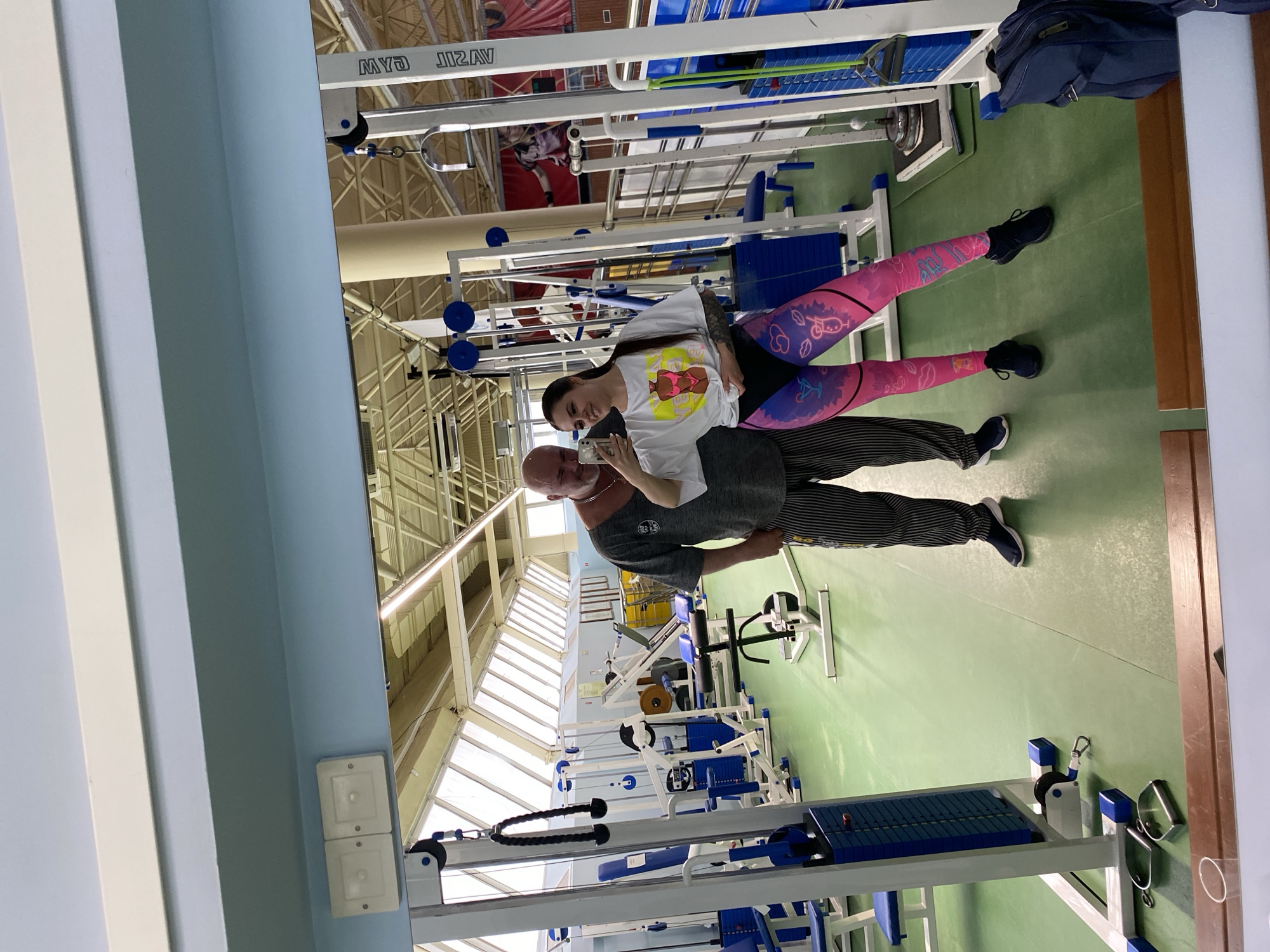 The image shows two people in a gym-like setting, likely in Chaikovsky, Russia. A woman is standing beside a man, and they appear to be taking a selfie. The woman has long dark hair, and is wearing a white t-shirt with a colorful cartoon print, and colorful leggings. The man is older, bald, wearing a gray shirt and horizontally striped pants. The background shows gym equipment including weight machines with blue padding and steel frames. The building has a high ceiling with a visible roof structure and large windows allowing natural light into the room. The floor is green and the walls are a light blue in color.
