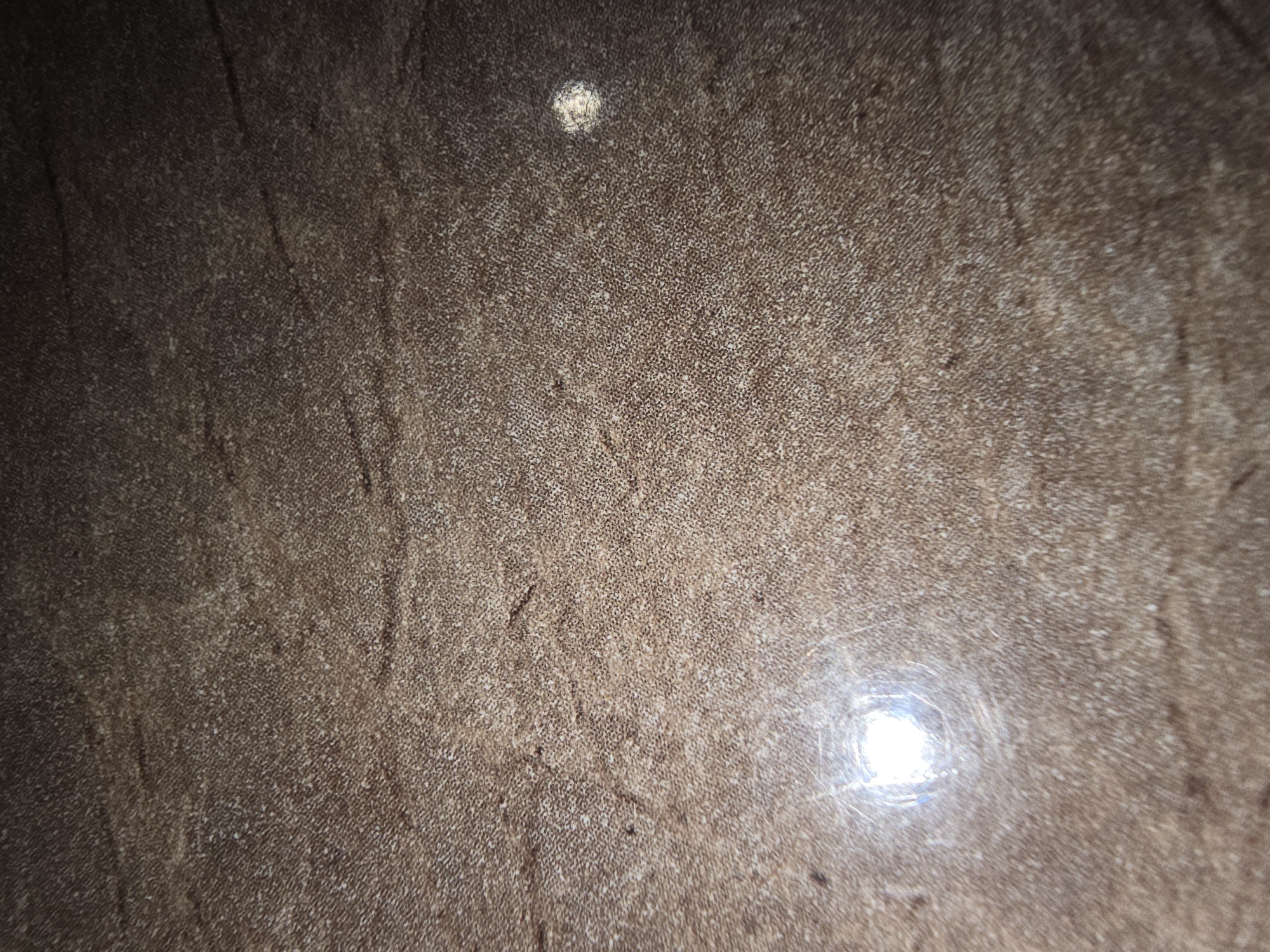 The image shows a close-up of a textured, brown surface that appears to be made of wood or a wood-like material. There are lighter specks and darker lines that create a wood grain pattern. Two distinct bright reflections are visible on the surface, suggesting it is smooth and possibly polished. One reflection is larger and more diffuse, located in the lower left quadrant, while the other is smaller and more concentrated in the upper right quadrant. The lighting suggests the image was taken in an indoor setting. There is no discernible location in the city of Ulan-Ude, Russia, as the image only depicts a surface. No people, objects, or actions are visible.