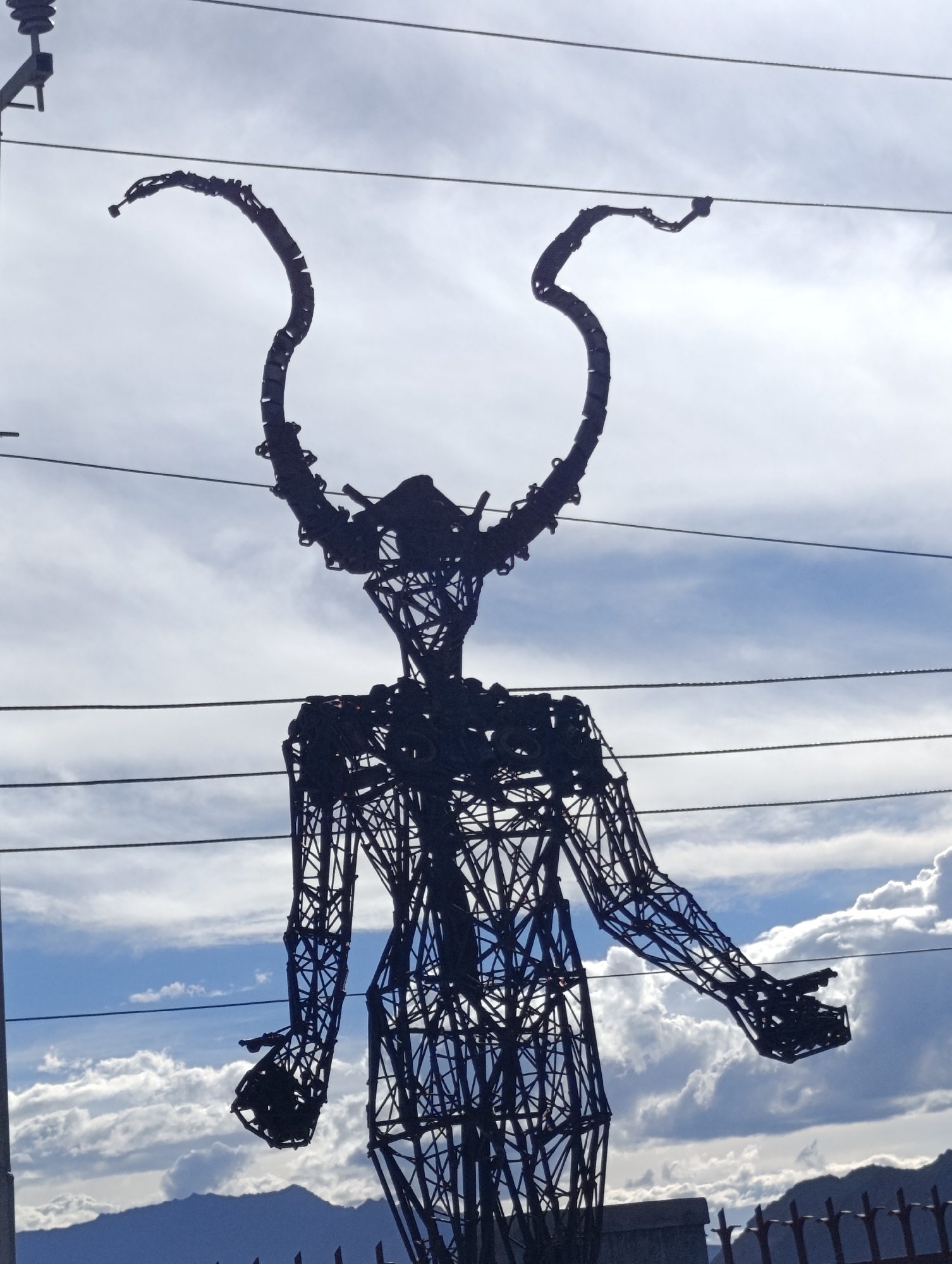 A large, dark, metal sculpture of a figure with outstretched arms and elaborate, curved horns stands silhouetted against a cloudy blue sky. The sculpture appears to be made of wire or thin metal rods forming a skeletal structure. It is positioned with multiple utility wires running horizontally in front of and behind it, suggesting an outdoor location, potentially within the city of Leh, India, given its mountainous backdrop and the characteristic utility poles and wires often found in such regions. The sculpture's pose, with arms extended and hands open, conveys a sense of welcome or offering. The lower portion of the sculpture is obscured by a dark, blurry object, possibly a wall or fence, and distant mountains are visible below the horizon line. The lighting suggests it is daytime, with sunlight filtering through the clouds, creating a dramatic contrast between the sculpture and the sky.