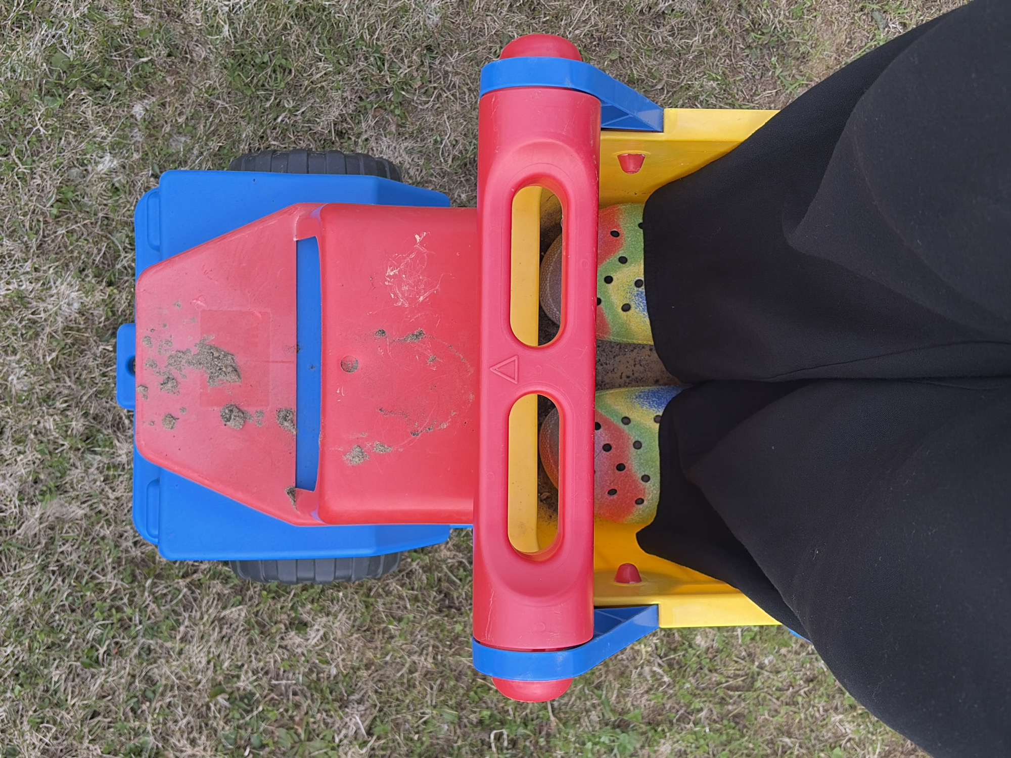 An overhead photograph captures a person seated on a brightly colored plastic ride-on toy outdoors in Chaikovsky, Russia. The toy is primarily red and blue, with a yellow footrest and seat area. The main red body of the toy is covered with patches of dirt and shows some wear. Blue elements form the base and parts of the steering/handlebar column, which is also red and features two elongated openings. Black wheels are visible on the left side of the toy.
Only the lower legs and feet of the person are visible, dressed in black pants. Their feet are resting on the yellow section of the toy, wearing vibrant, multi-colored clogs patterned with green, yellow, red, and blue dots.
The setting appears to be a yard or open space with a ground cover of sparse, dry grass and visible dirt. The natural lighting suggests it is daytime. The scene depicts a moment of play or rest on the toy. No visible text is present in the image.