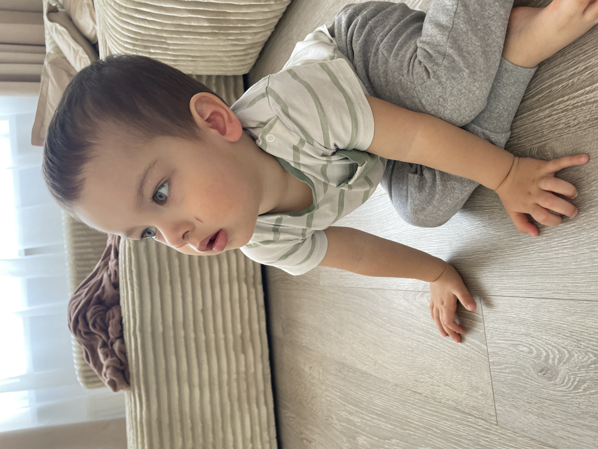 A toddler lies prone on a light-colored wooden floor next to a couch with light tan cushions. The child wears grey sweatpants and a light-colored striped shirt. The toddler's left cheek has a minor scratch. The location cannot be determined to be in Izhevsk, Russia based on the image content.