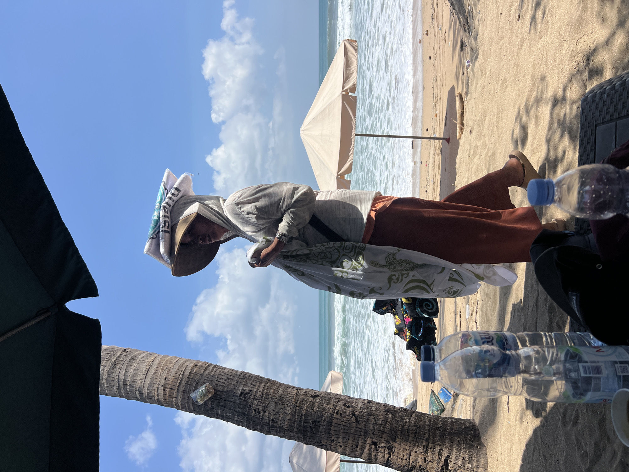 A person wearing a sun hat and a hooded garment stands on a sandy beach in Kuta, Indonesia, carrying a stack of items on their head and a large patterned fabric. The person is also holding a smaller fabric in their hands. In the foreground, several plastic water bottles are visible on the sand, along with a dark object that appears to be a bag or backpack. A palm tree trunk is on the left side of the frame, and a dark umbrella extends from the top. In the background, the ocean with waves is visible under a bright blue sky with scattered clouds. Two beach umbrellas are set up on the sand near the water's edge. The scene depicts a sunny day at the beach.