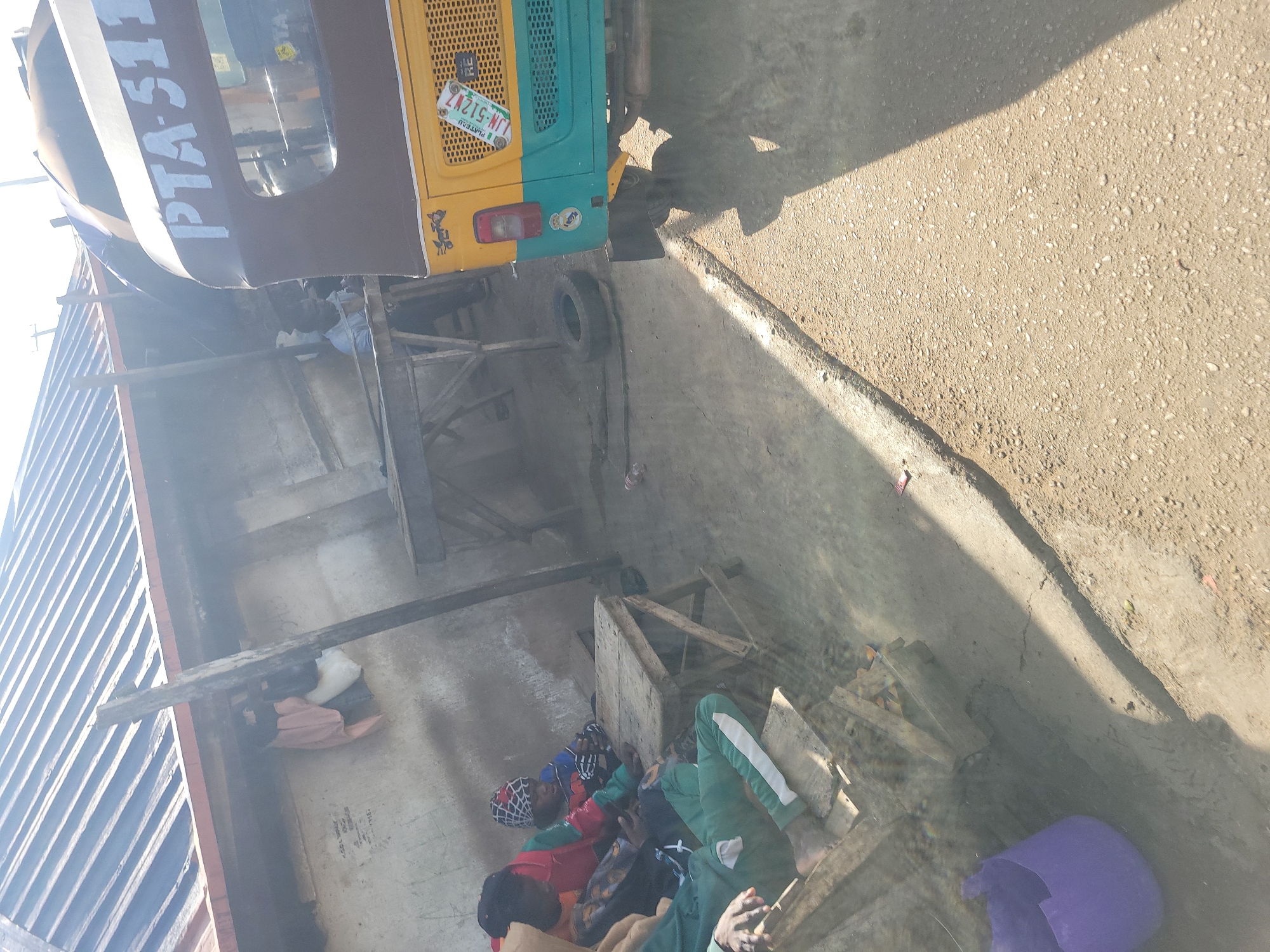 The image depicts a street scene in Jos, Nigeria, with a focus on a roadside area adjacent to a building with a corrugated metal roof. In the foreground, a yellow and teal motorized tricycle, possibly a tuk-tuk, is parked. The tricycle has a license plate that reads "LJN 512 W7 RE PLATEAU". On the side of the tricycle, a white inscription reads "PTA-511".
To the left of the tricycle, a group of people, including children, are seated on wooden structures or benches. They appear to be gathered around a small wooden table. One person is wearing green trousers with white stripes. Another individual is wearing a head covering with a patterned design. The building behind them has a weathered, concrete-like wall. Hanging from an overhead beam of the building are some pieces of clothing.
Further back, there's a wooden structure that resembles a market stall or workshop. A spare tire is visible on the ground near this structure. The ground is unpaved and appears dusty, with some debris scattered around. The overall environment suggests an informal urban setting. The lighting indicates it is daytime, with strong sunlight casting shadows.