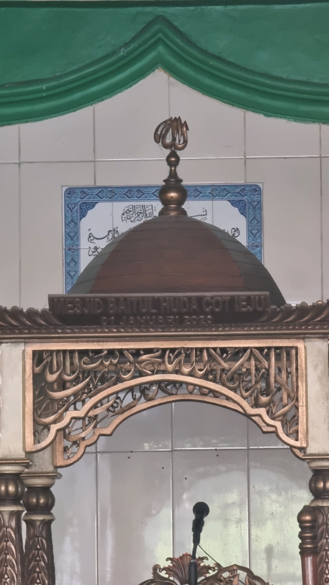 The image shows an ornate wooden and metallic structure, possibly a pulpit or a decorative element within a mosque. A dome-shaped structure is visible at the top, topped with a metallic ornament that appears to be Arabic calligraphy. Below the dome, text is inscribed on a horizontal band, reading "MESJID BAITUL HUDA COT IEJU" and "01 JANUARI 2020". The structure is adorned with intricate Arabic calligraphy carved into a golden archway. In the background, light-colored tiles cover the wall, and a green draped decoration is visible at the top. A black microphone is positioned in front of the structure, suggesting it is a functional element for speeches or prayers. The overall impression is one of religious and cultural significance, likely within a mosque in Blangrambong, Indonesia, given the inscription on the structure.
