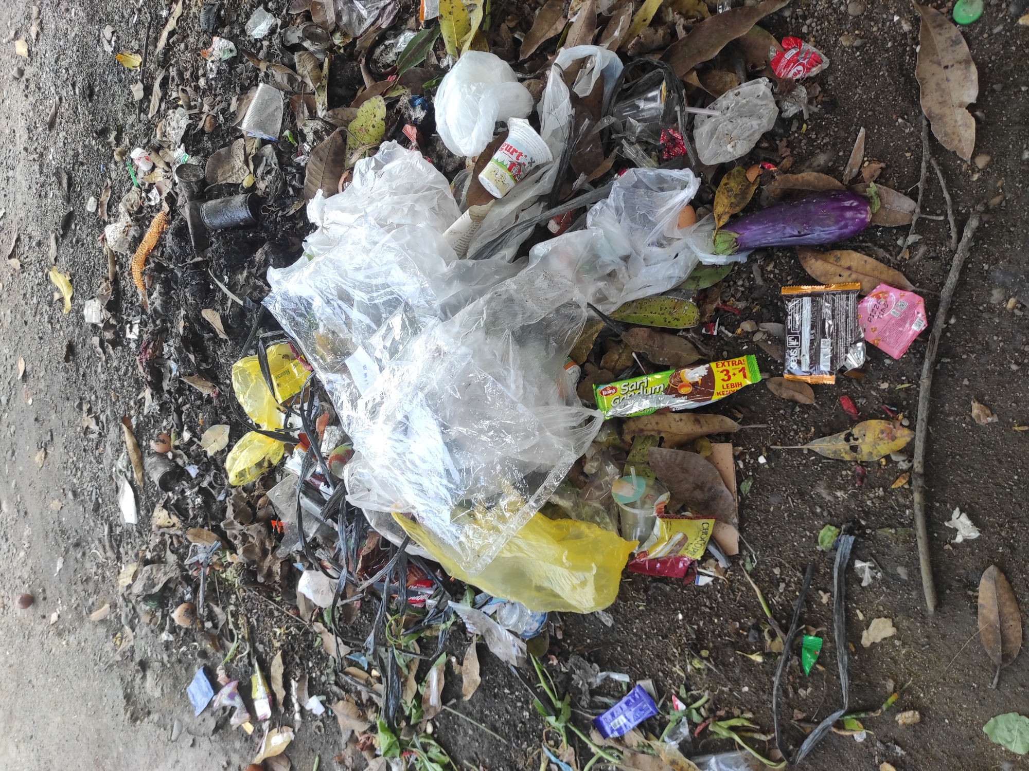 The image captures an uncontrolled accumulation of refuse on an unpaved ground surface in Lhokseumawe, Indonesia. The ground comprises dark soil and gravel, interspersed with scattered dry leaves and sparse green vegetation. A substantial pile of mixed waste occupies the central and lower portions of the frame. Identified items within the waste include multiple transparent plastic bags, several yellow plastic bags, and various plastic food wrappers, such as those for "SARI GANDUM" chocolate, "CHOC O' PIE", a "SNICKERS"-like bar, and a pink "PIZZA TIME" wrapper. Also visible are plastic beverage cups, one with the text "Yumm!", and a red wrapper from "YAYAYA". Organic waste elements include a solitary purple eggplant, a brown egg, and numerous dried leaves and small branches. Several dark, potentially charred, metal cans are embedded within the debris, alongside tangled black wires or cables. A piece of orange, porous material resembling honeycomb is also present. The ground exhibits patches of darkened, possibly burnt, earth. No individuals are depicted in the scene.