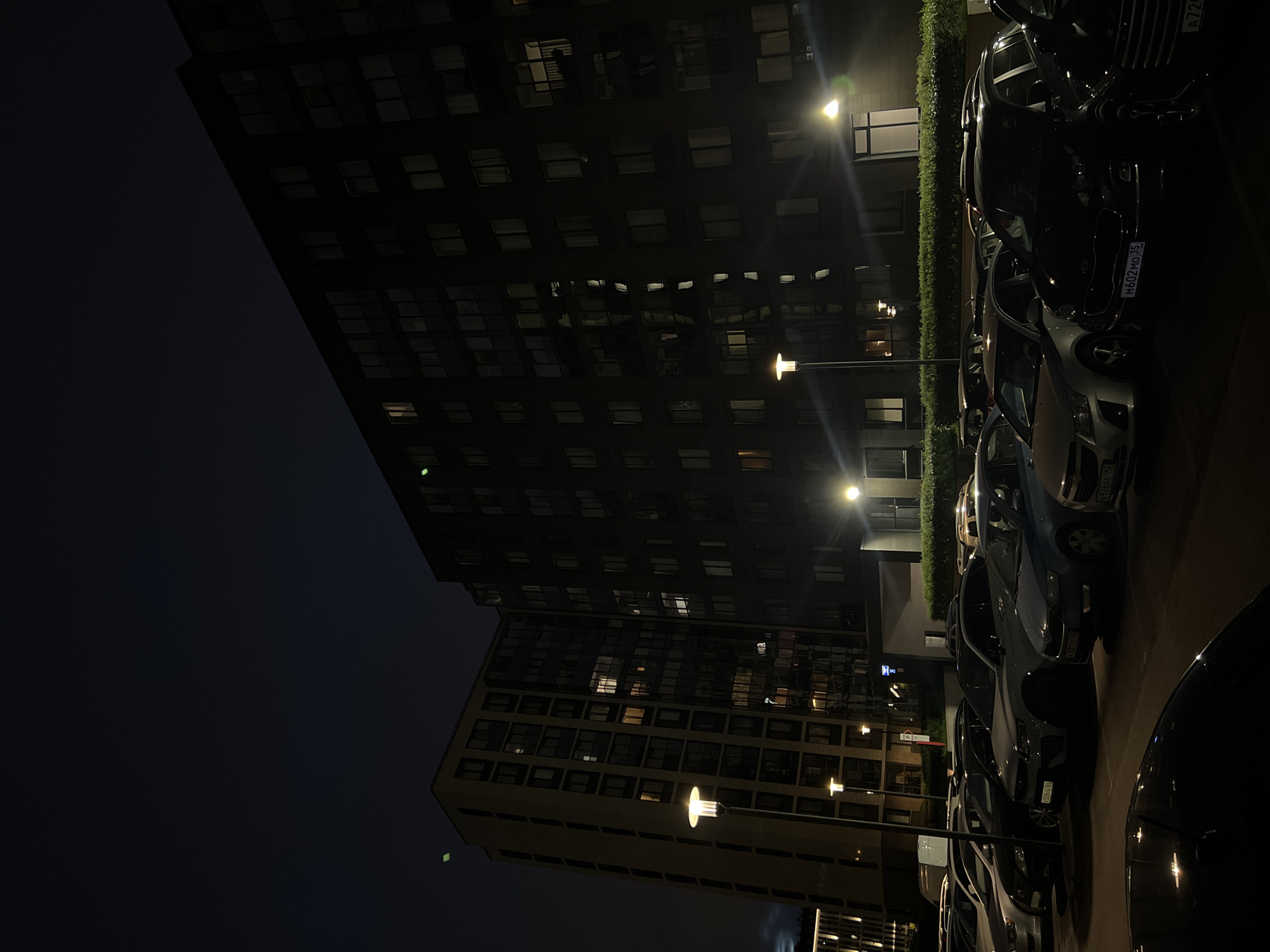 An outdoor nighttime scene in an urban environment depicts a parking lot filled with multiple vehicles parked in front of two multi-story apartment buildings. The buildings are dark, with some windows illuminated, indicating occupancy. Streetlights are visible, casting a warm glow on the pavement and illuminating the parked cars. The sky above is dark and appears to be overcast or heavily polluted, obscuring any celestial bodies. The overall impression is one of a quiet, residential area at night.